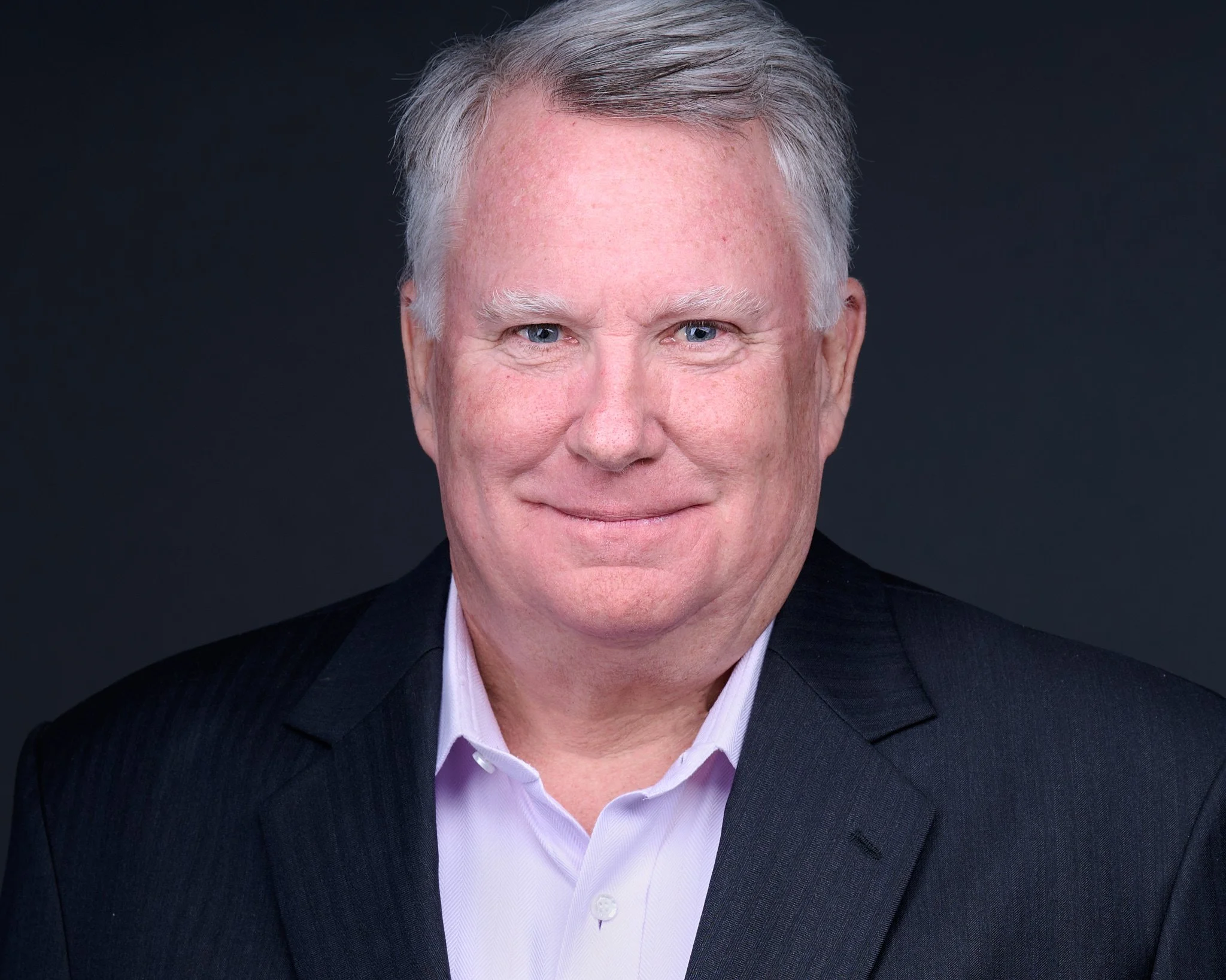 A headshot of a middle-aged man with gray hair, blue eyes, and fair skin, wearing a dark suit jacket and a white dress shirt against a dark background.