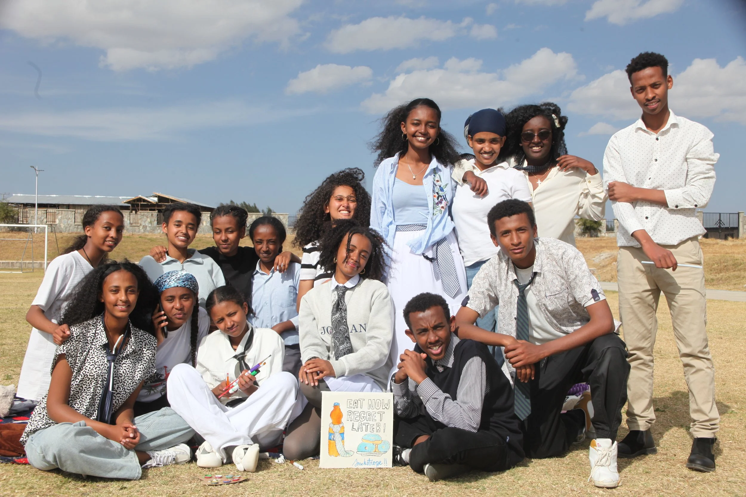 A group of young students and their teacher posing outdoors on a sunny day with blue sky and clouds, holding a sign that says, 'Eat Now Regret Later!'