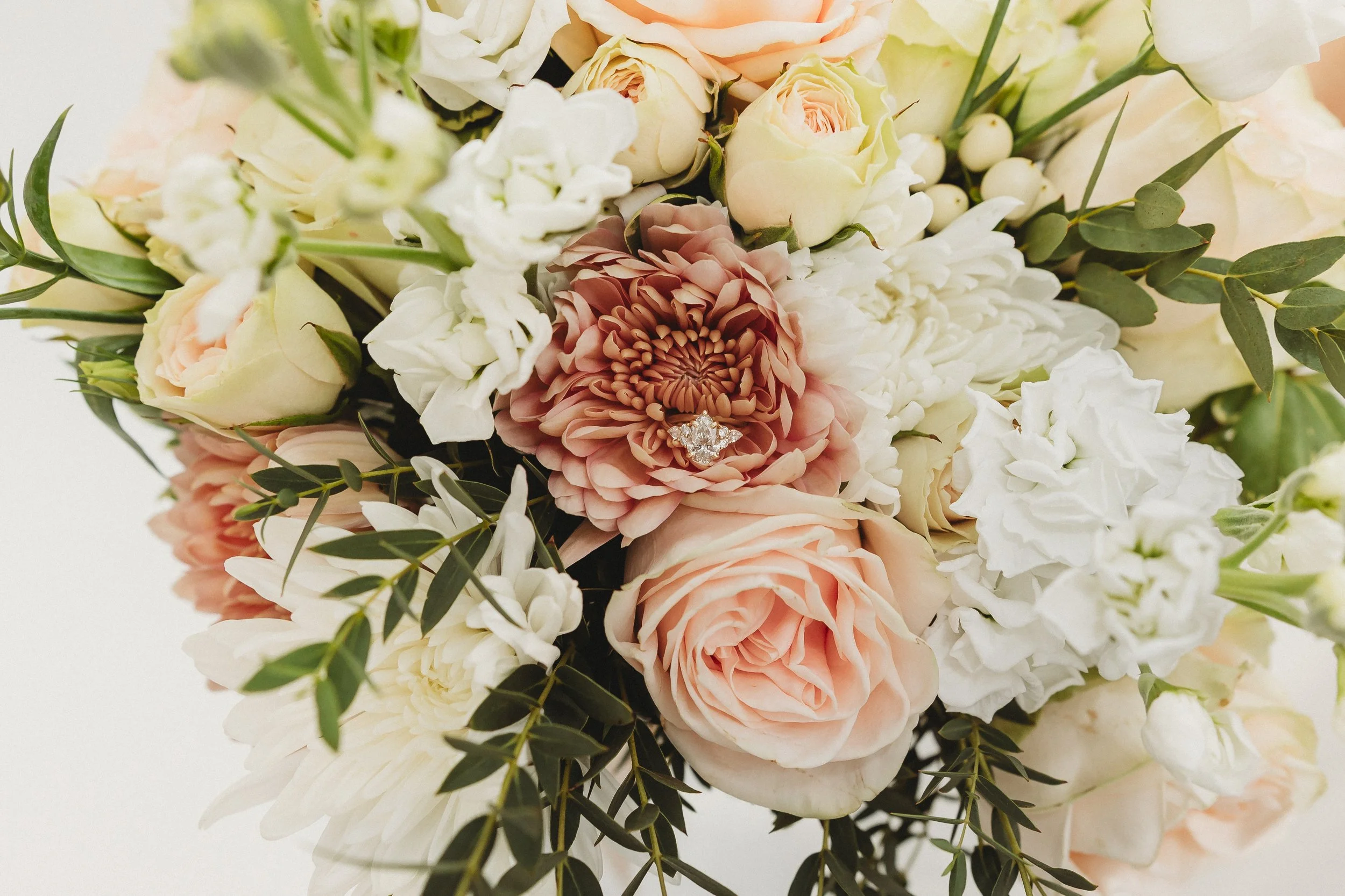 Close-up of a wedding bouquet with white, peach, and blush roses, hydrangeas, and greenery, with an engagement ring placed on a large peach dahlia.