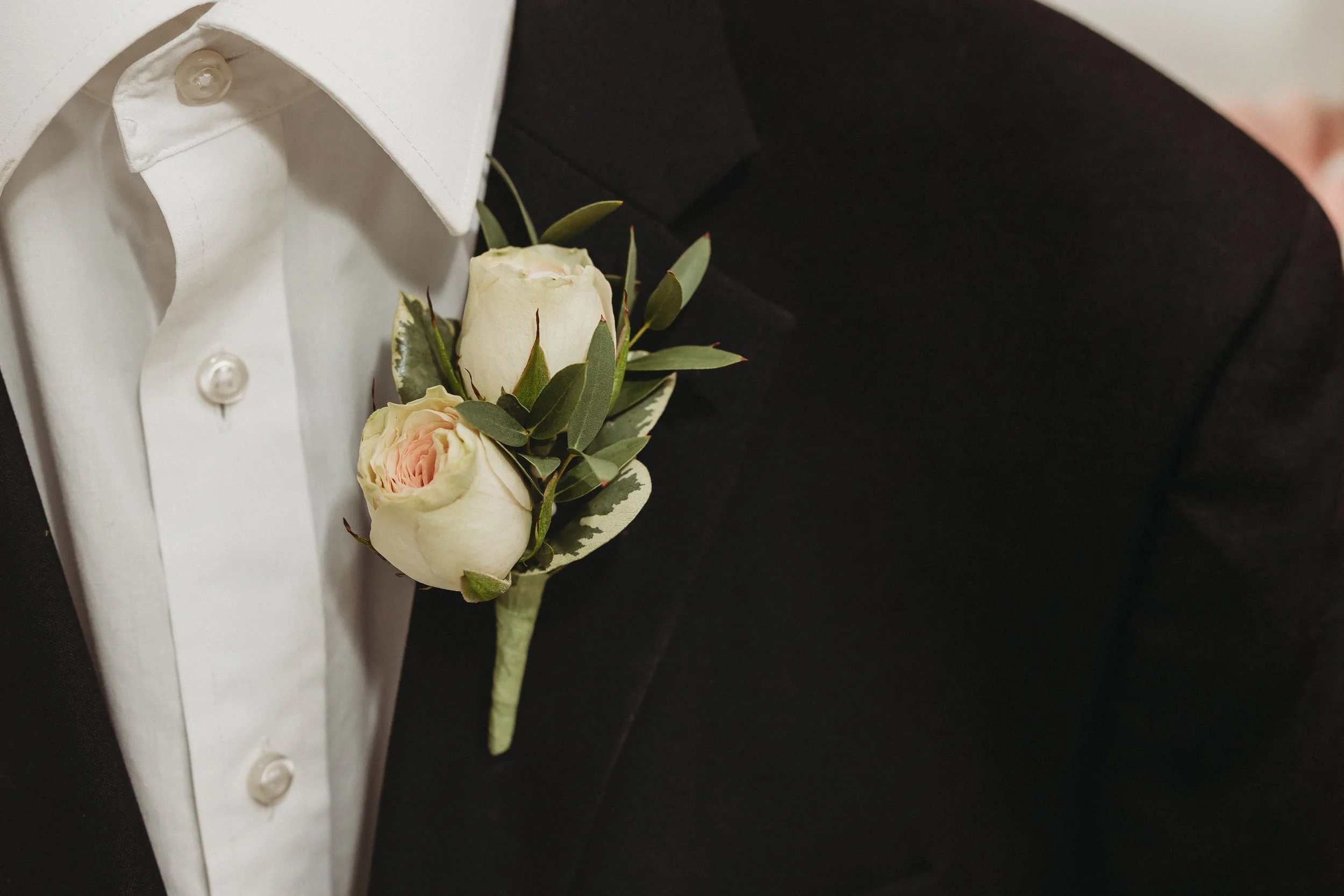 White boutonniere with green leaves pinned to the white shirt of a black suit. This boutonniere is in the Cloud Nine collection palette.
