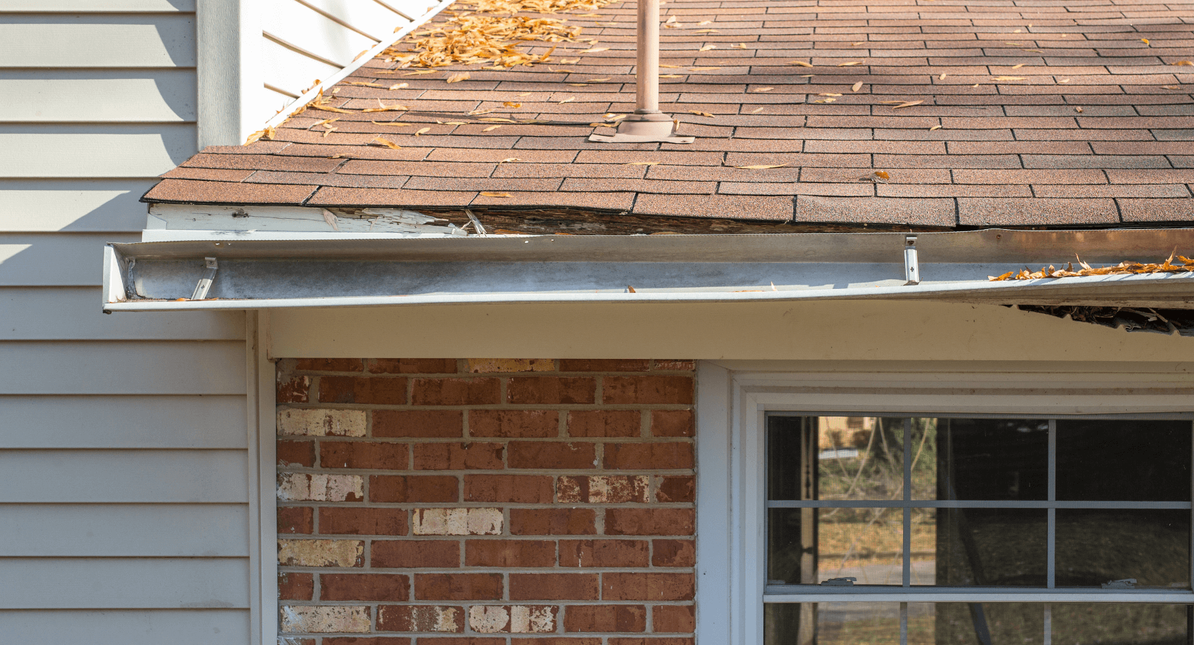Close-up view of a house's roof and exterior wall showing damage. The roof has missing shingles, debris, and a damaged gutter with leaves inside. The house features a brick wall next to white vinyl siding and a window.