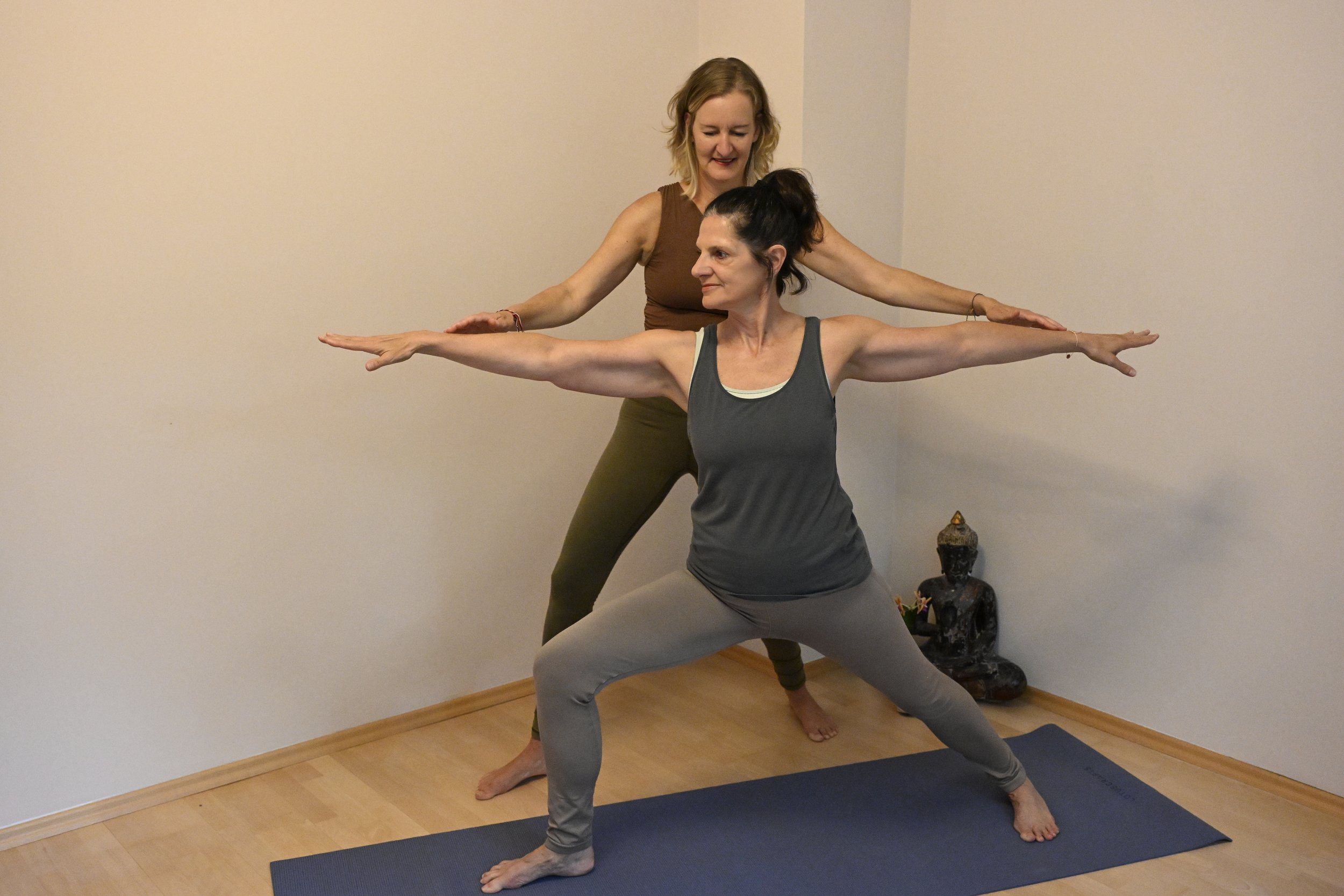 Zwei Frauen beim Yoga, eine in der Pose des Kriegers mit weit ausgestreckten Armen, die andere dahinter, die Anweisungen gibt.