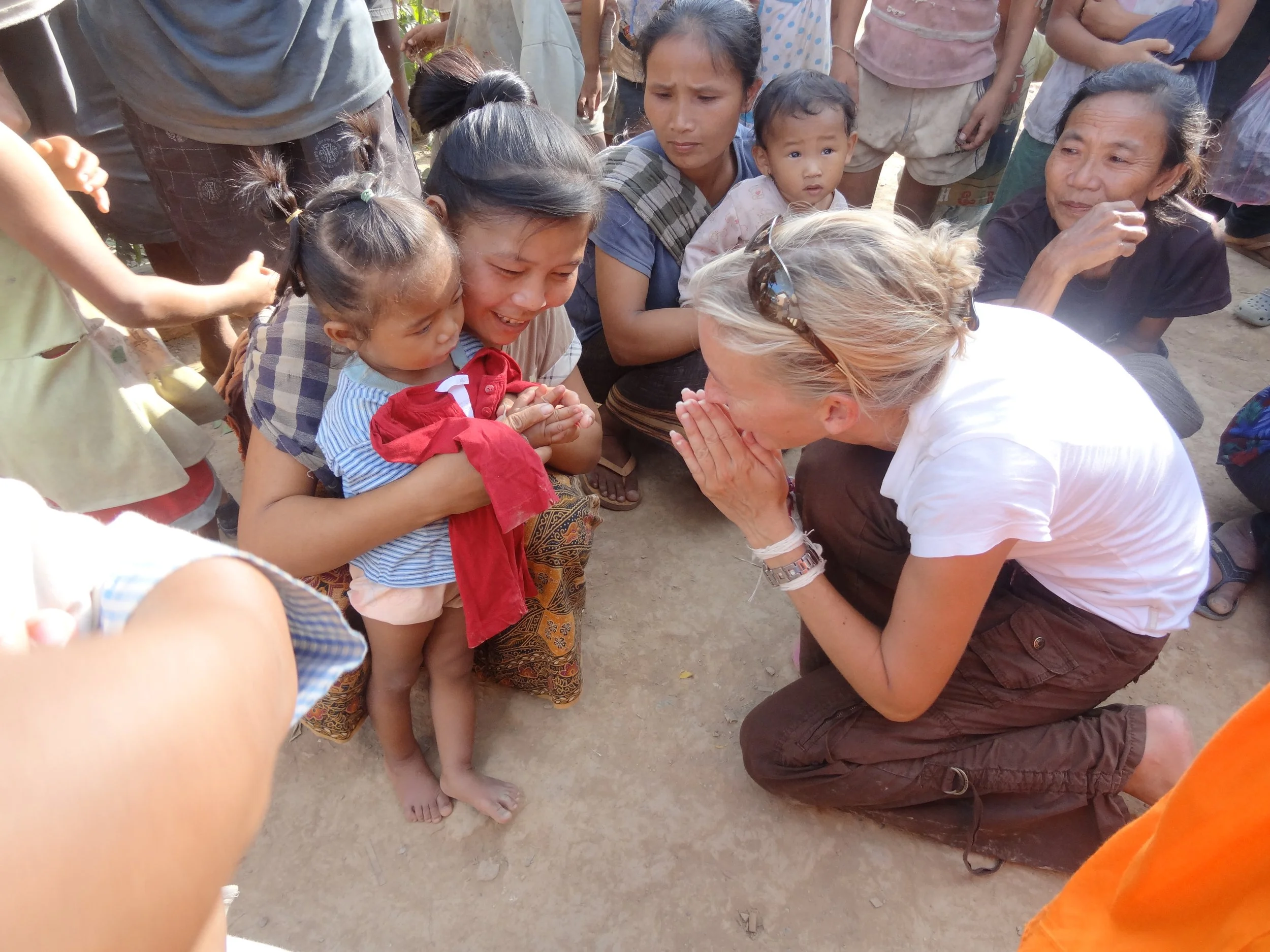 Eine blonde Frau kniet auf dem Boden und begrüßt eine junge asiatische Mädchenfamilie mit gefalteten Händen und einem Lächeln. Die Szene findet im Freien statt, umgeben von mehreren anderen Kindern und Erwachsenen.