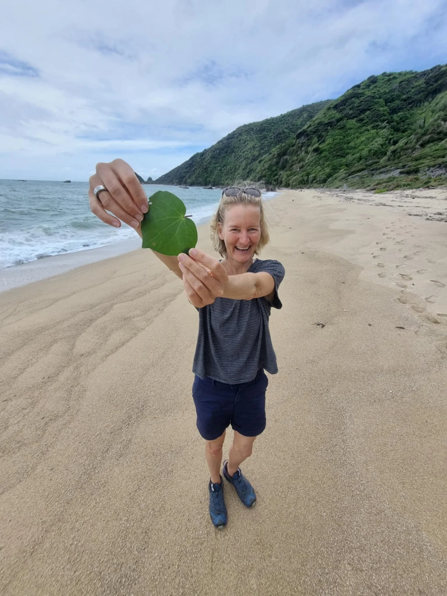 Frau am Strand hält ein grünes Blatt in die Kamera, lächelt und zeigt es stolz. Im Hintergrund sandiger Strand, Meer und grüne Hügel bei bewölktem Himmel.