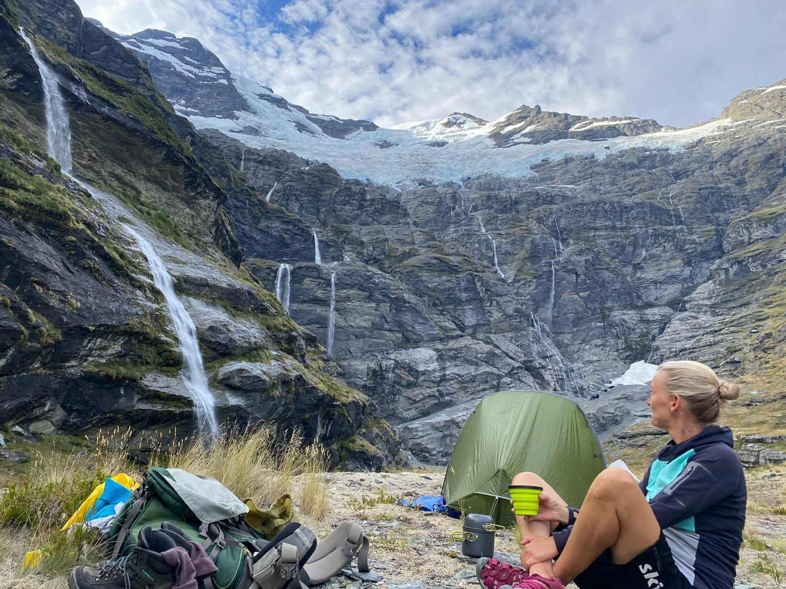 Frau sitzt vor einem Zelt in einer Berglandschaft mit Wasserfällen und Gletschern, hält eine grüne Tasse, umgeben von Wanderausrüstung.