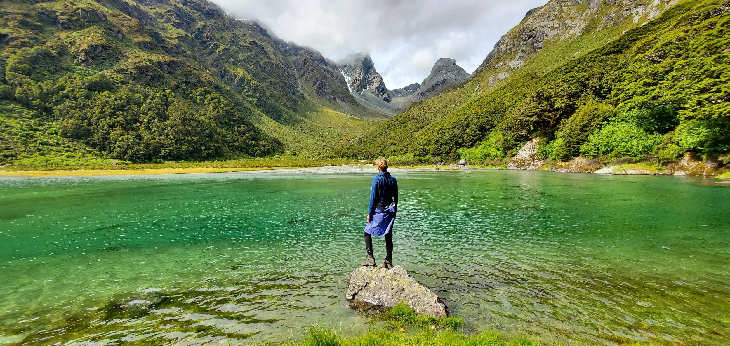 Person steht auf einem Felsen am Ufer eines grünen Sees, umgeben von grünen Bergen und Wolken am Himmel.
