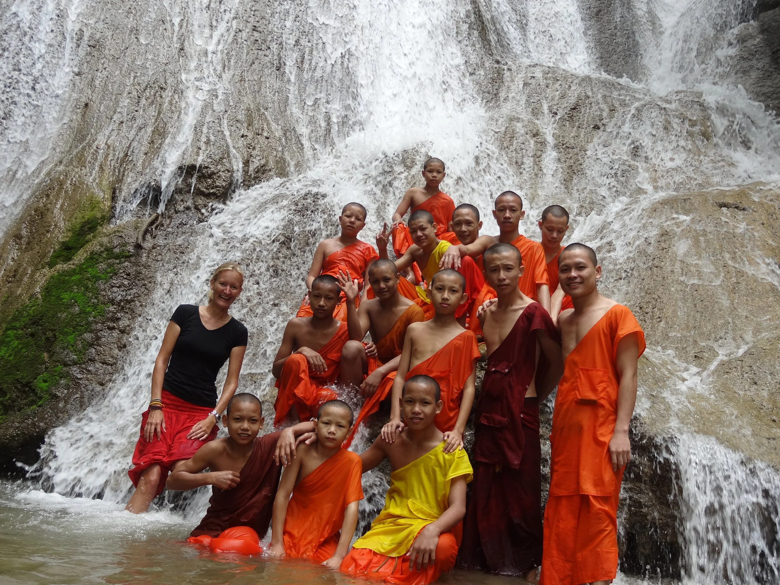 Eine Gruppe von buddhistischen Mönchen in Orange und braun, zusammen mit einer deutschen Frau, posieren vor einem Wasserfall.