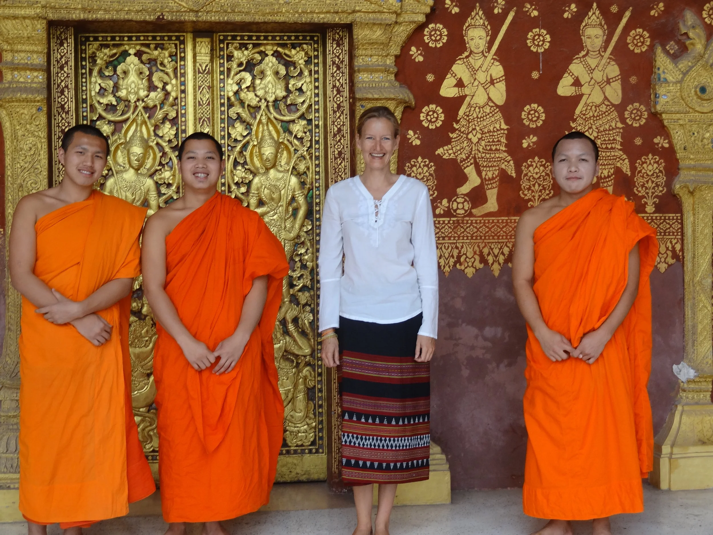 Vier Menschen stehen vor einer goldverzierten Wand. Drei junge buddhistische Mönche tragen orange Roben, eine Frau trägt eine weiße Bluse und ein langes, bunt gemustertes Rock. Alle lächeln.