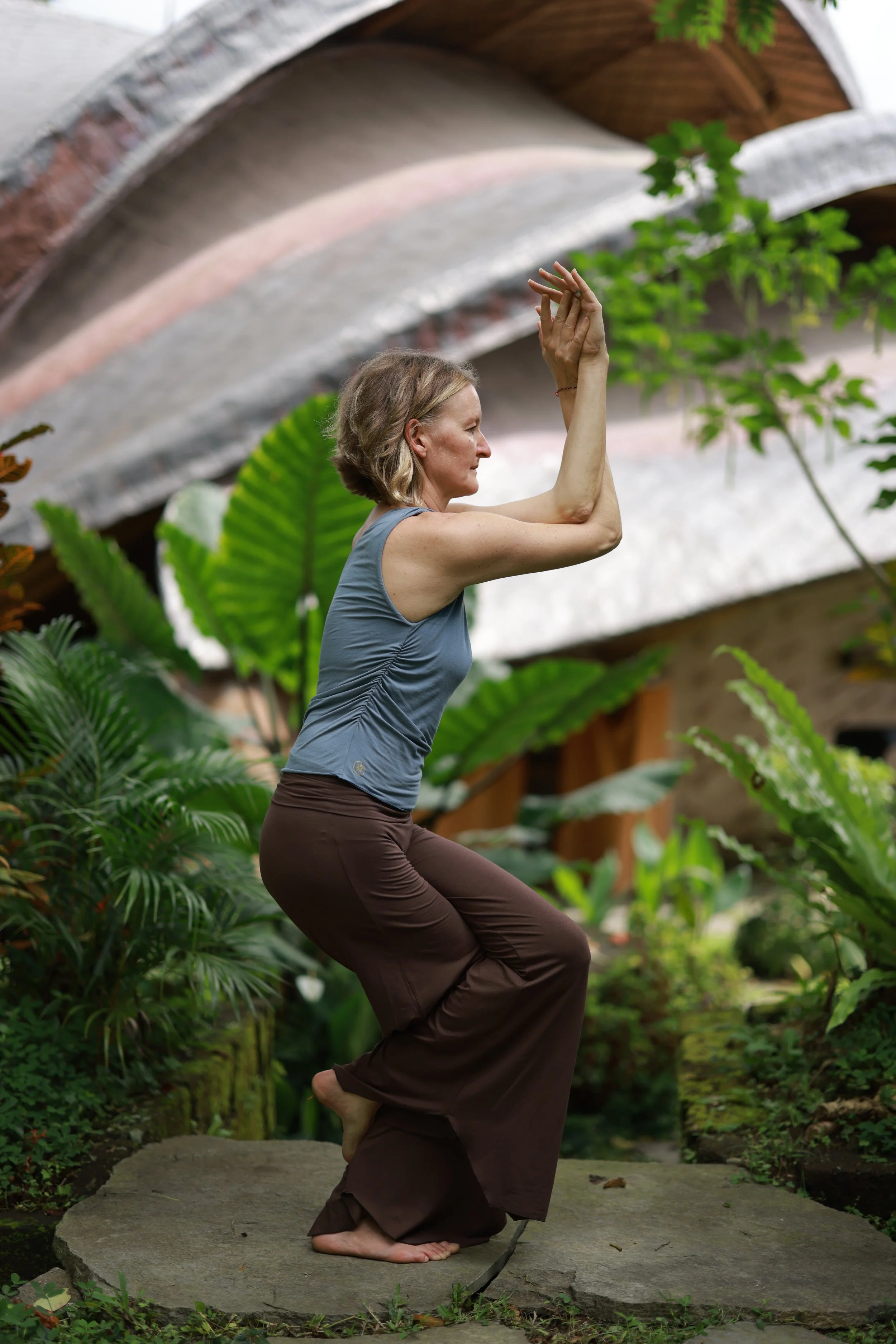 Eine Frau praktiziert Yoga in einem grünen Garten, sitzt auf einem Stein und hebt die Arme über den Kopf. Sie trägt eine ärmellose graue Sportjacke und braune Hosen.