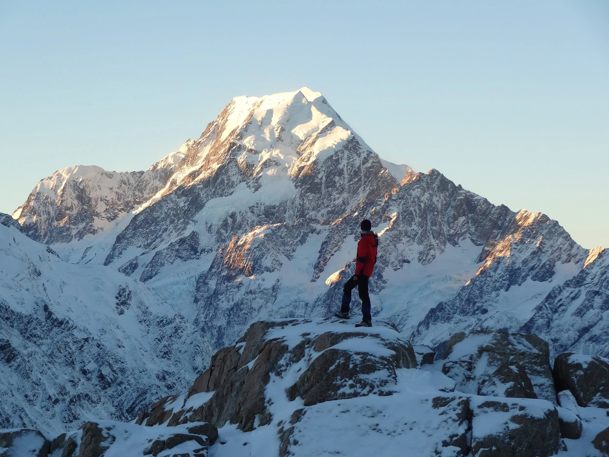Ein Mensch in einer roten Jacke und schwarzen Hosen steht auf felsigen, schneebedeckten Terrain vor einer großen, schneebedeckten Bergkette.
