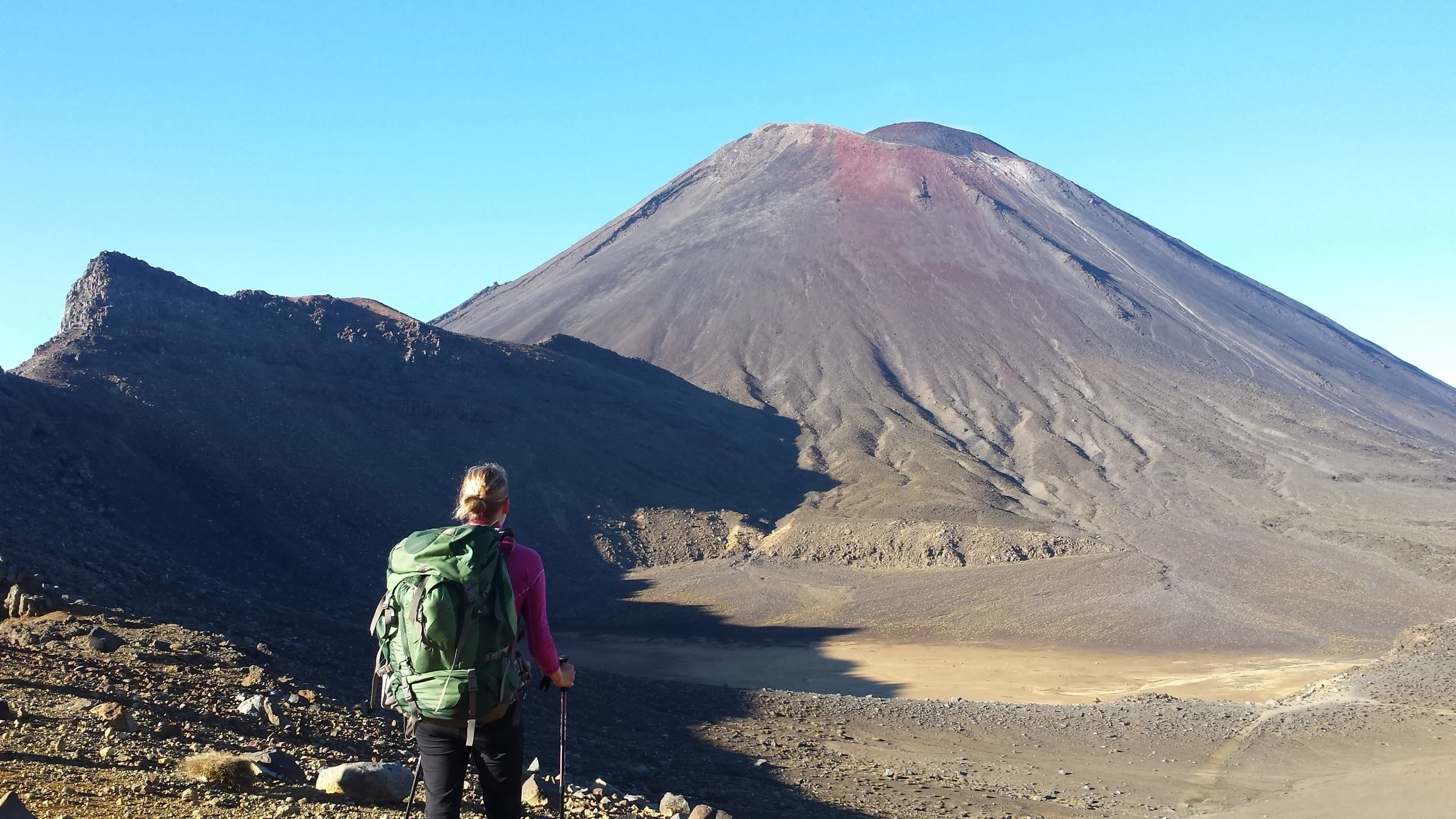 Eine Person mit Rucksack und Wanderstöcken schaut auf einen Vulkan bei klarem Himmel.