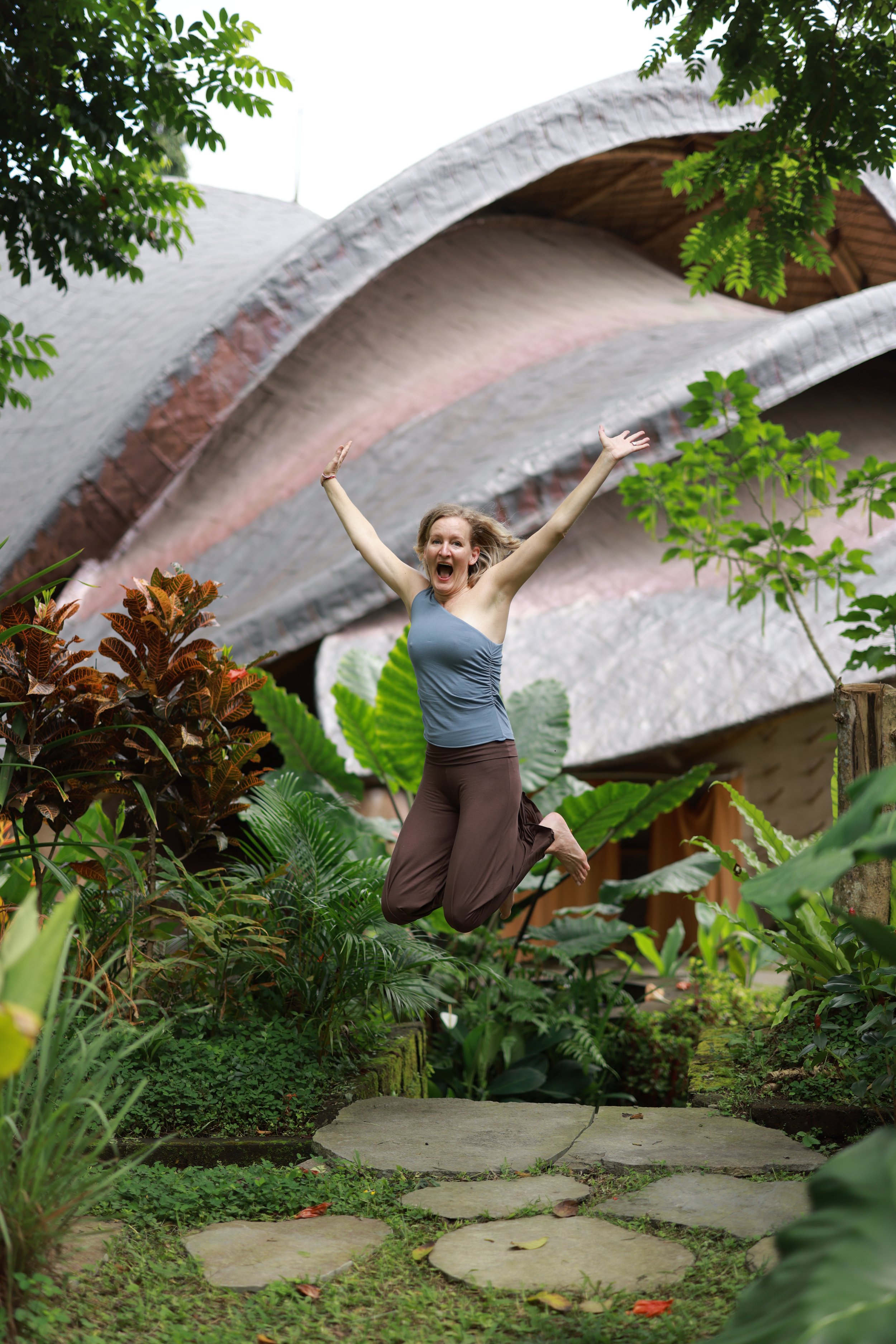 Frau jubelt springt in einem Garten mit tropischen Pflanzen und modernen Gebäuden im Hintergrund.