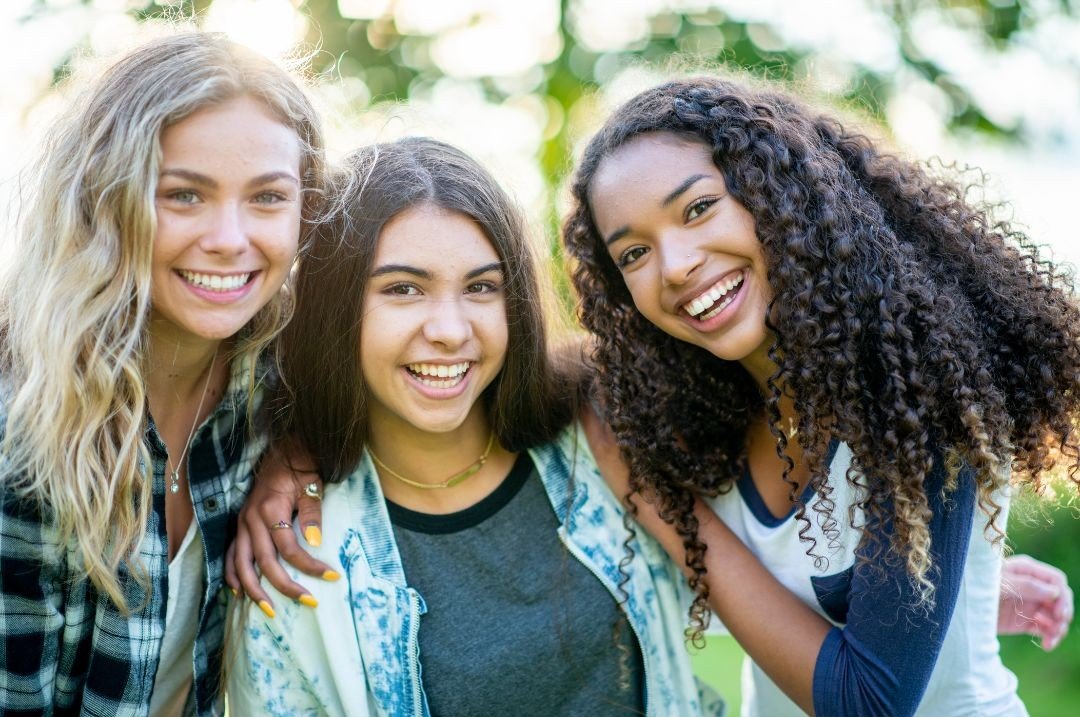 Three young women smiling and posing for a photo outdoors, with greenery in the background.