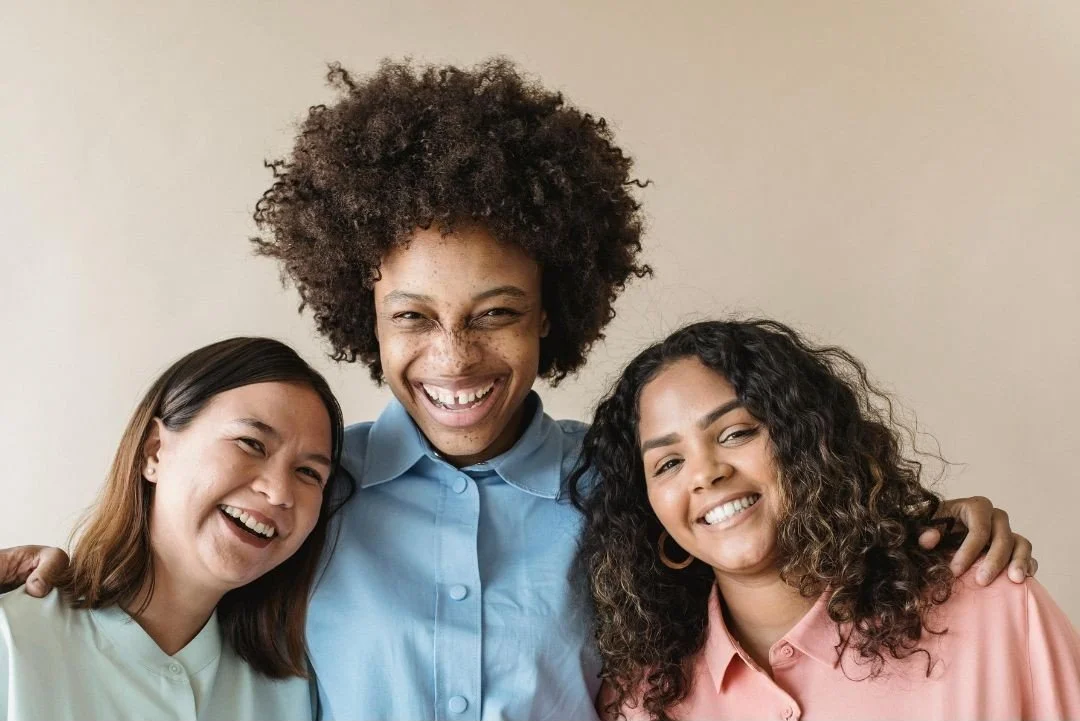 Three women smiling and posing together indoors, with one woman in the center having curly hair and wearing a blue shirt, and two women on either side with straight and curly hair, wearing pastel-colored tops.