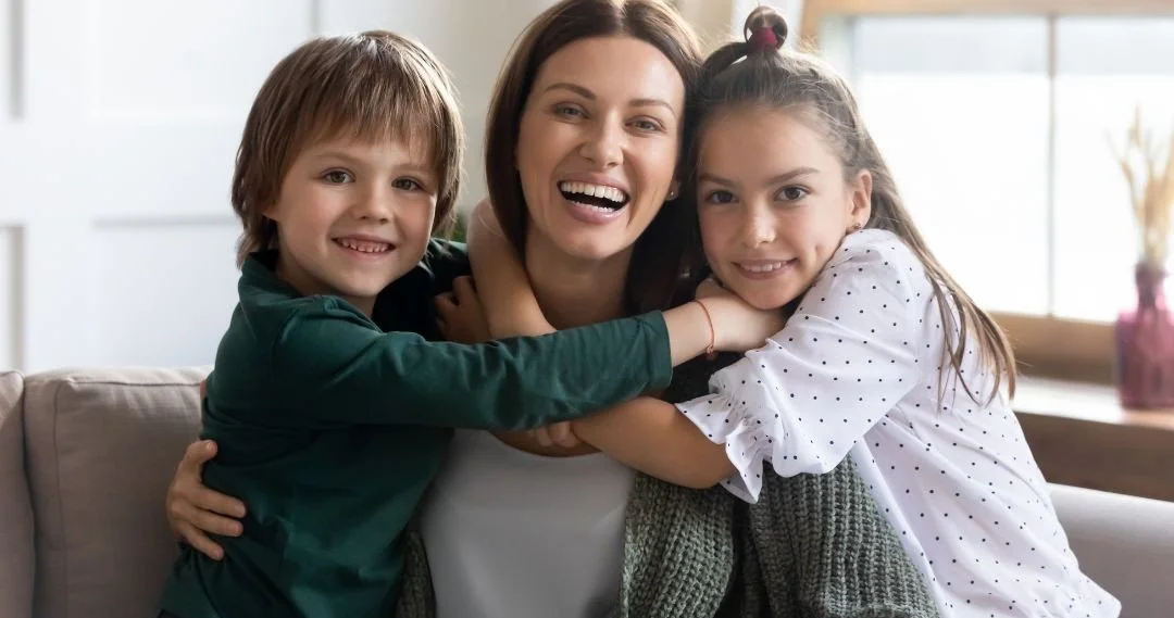 A woman with two children hugging and smiling on a couch in a bright living room.