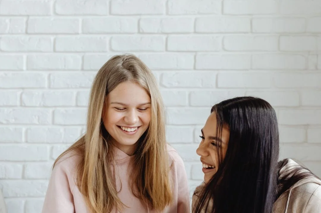 Two young teens smiling and chatting together in front of a white brick wall