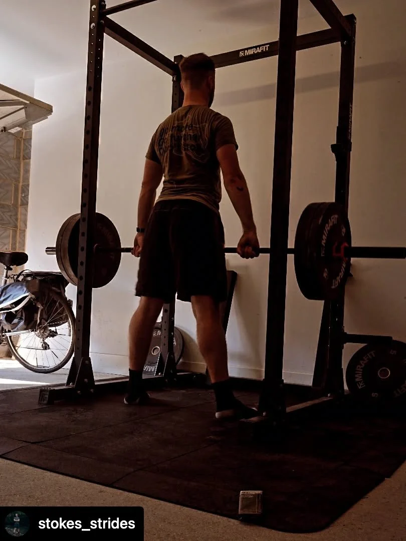 A man lifting weights with a barbell in a home gym, standing on a black mat, near a bicycle, with a squat rack labeled 'MIRAFIT'.