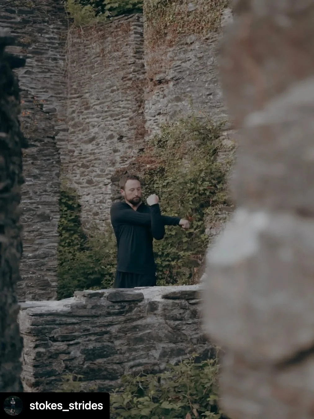 A man in black workout clothes stretching his arm against a stone wall in an outdoor setting with old stone ruins and green foliage.