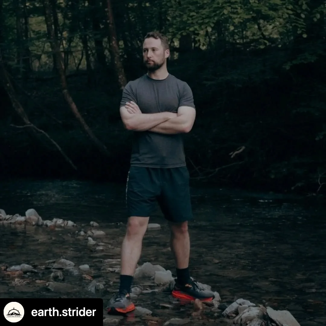 A man standing with arms crossed on a rocky riverbank in a forested area, wearing a dark athletic shirt, shorts, and trail running shoes.