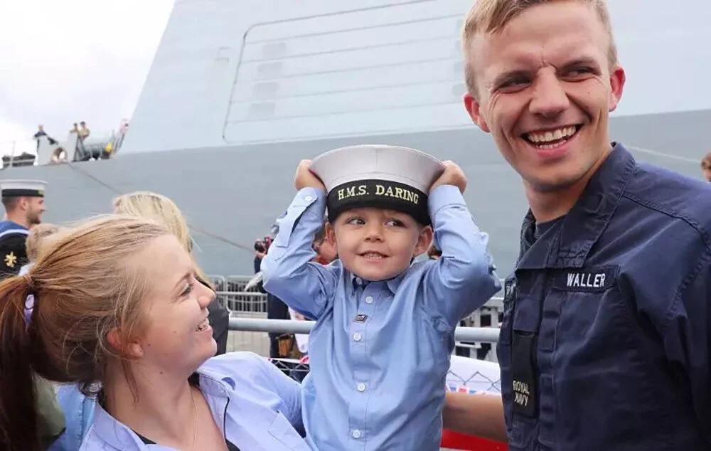 A family of three celebrating at a naval event, with a large gray ship in the background. The young boy is wearing a naval cap that says 'H.M.S. DARING' and is being held by a woman while smiling. The man, wearing a navy uniform with a name tag 'WALLER', is smiling at the camera.