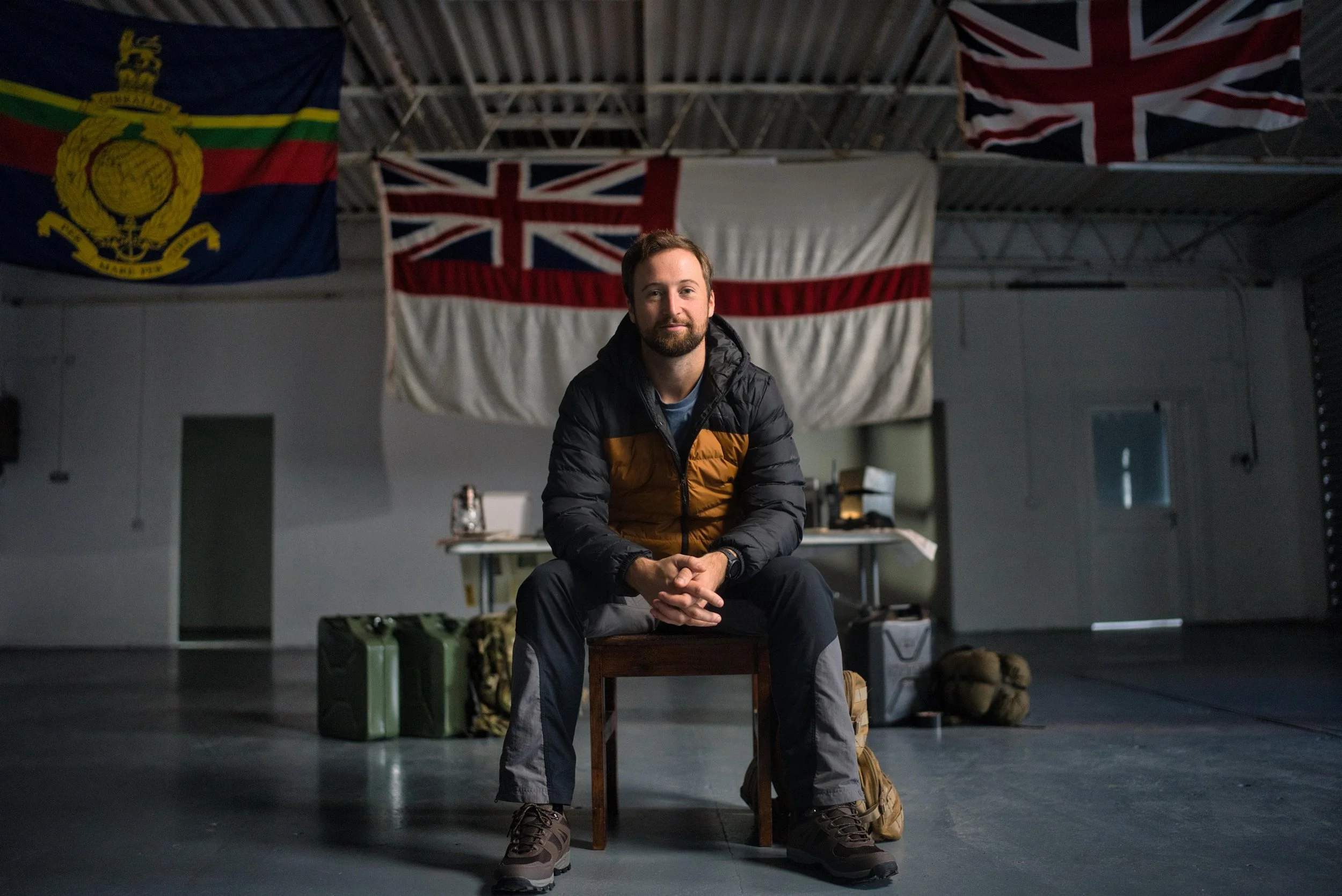 Fred Stokes sitting on a chair inside a room with flags hanging on the wall, including the Union Jack, and military gear and supplies on the floor.