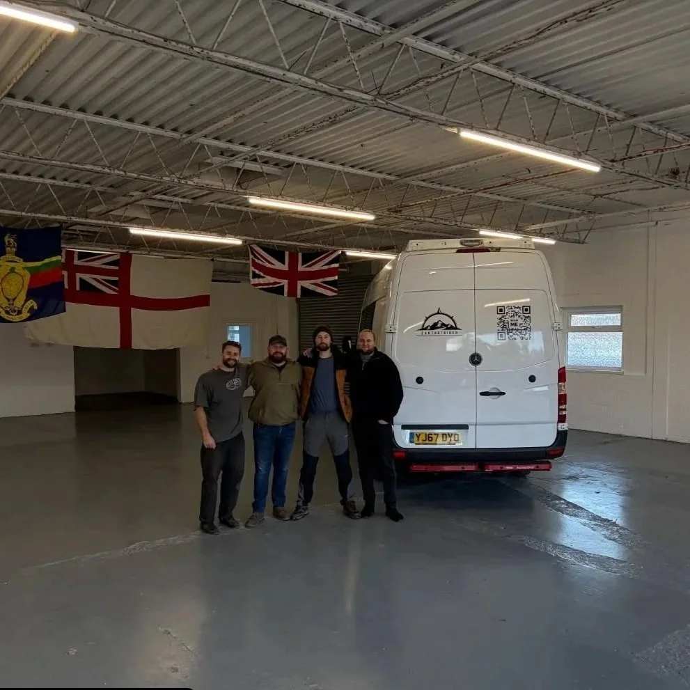Four men standing together in a warehouse-like space with flags hanging on the wall and a white van behind them. The flags include the Union Jack and the English flag.