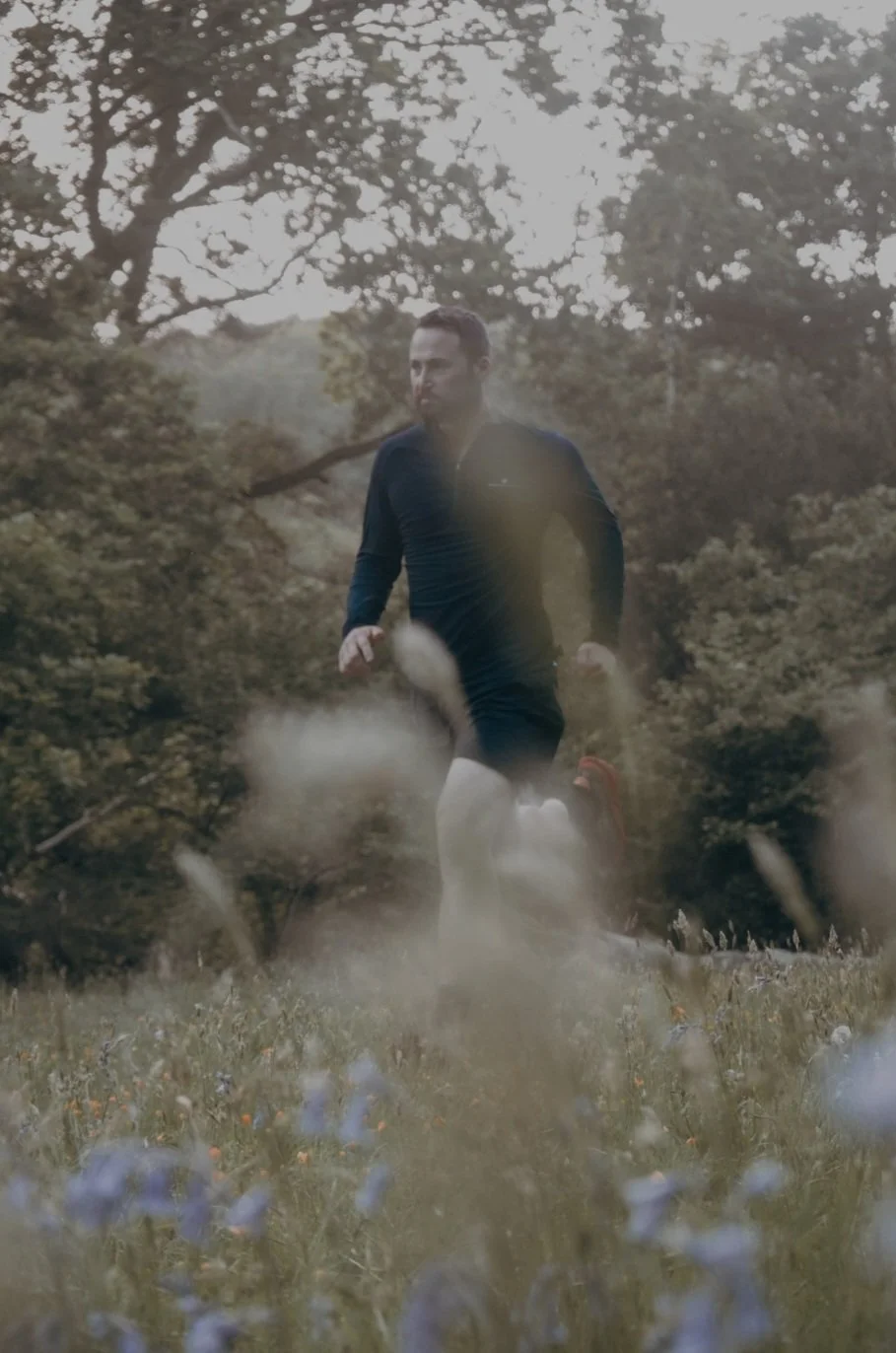 A man running through a grassy field with wildflowers, surrounded by trees and foliage, wearing a dark athletic jacket and shorts.