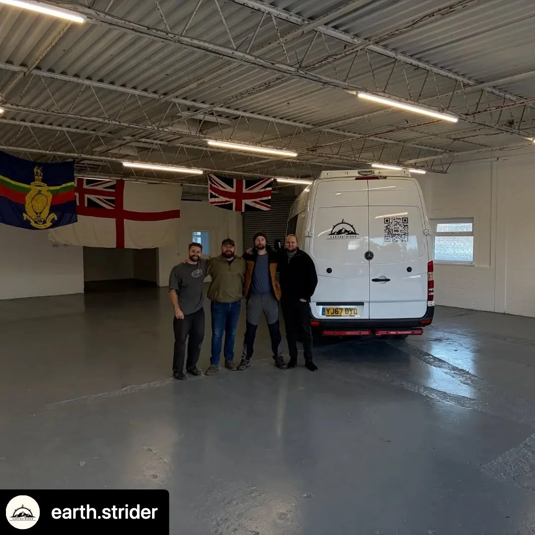 Four men stand inside a garage next to a white van with a mountain and QR code sticker on the back. Four flags hang on the wall behind them, including the Union Jack, England flag, and a flag with a crown and tricolor stripes. The garage has a high ceiling with overhead lighting.