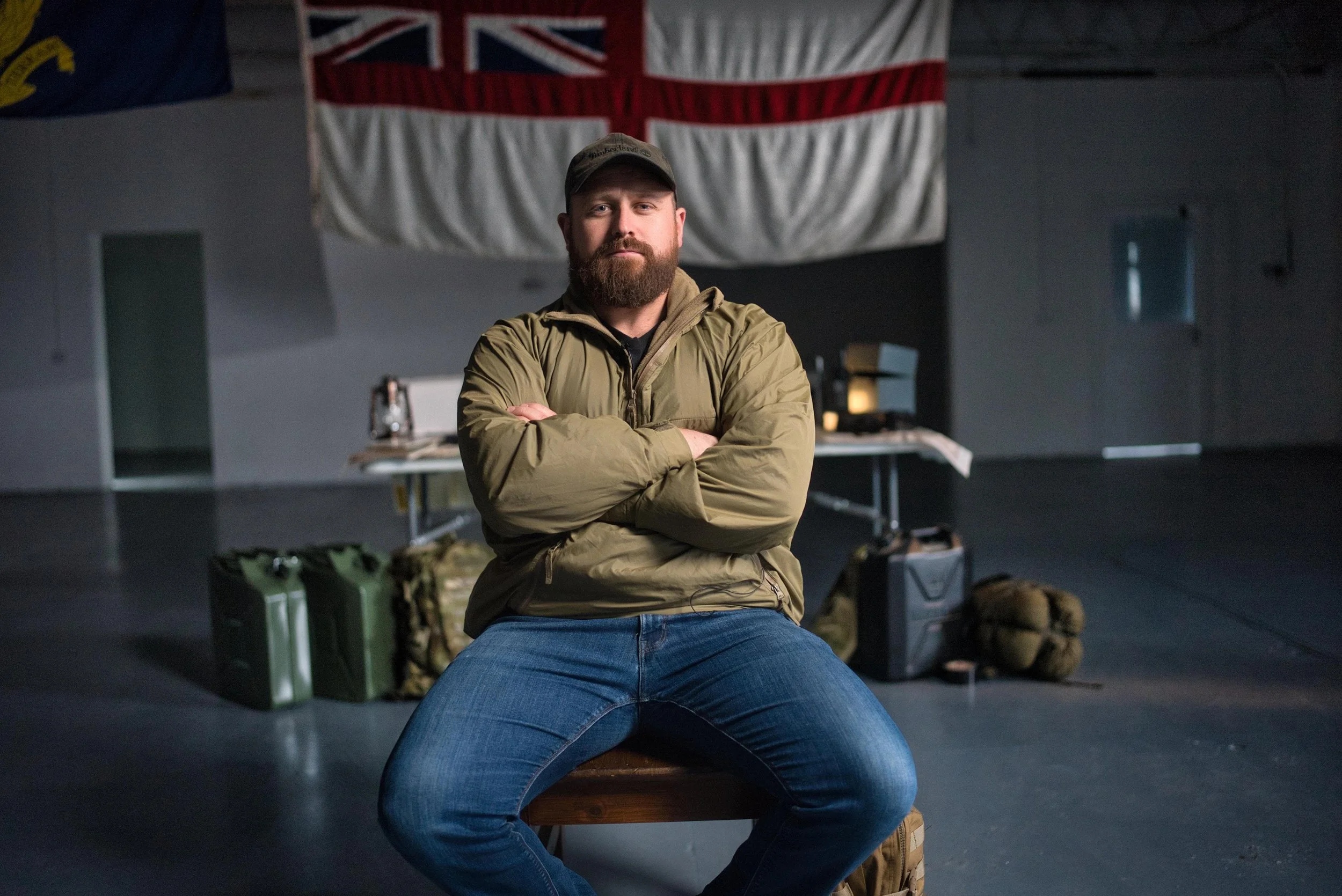 A man with a beard and wearing a cap, khaki jacket, and jeans sitting on a wooden bench with arms crossed inside a large indoor space. Behind him are left military-style containers, a camouflage backpack, and other gear. Large national flags are hanging from the ceiling, including the Union Jack.