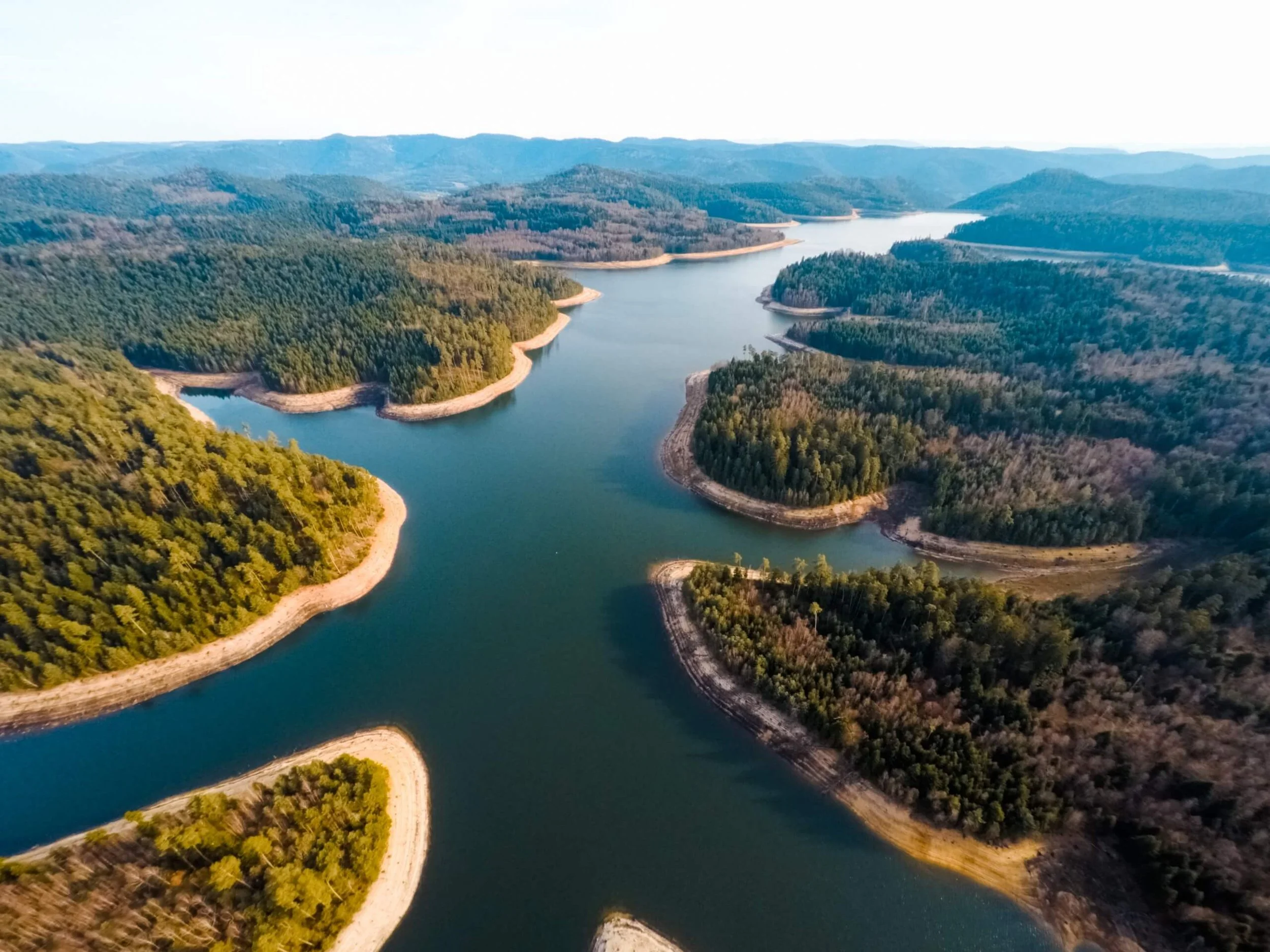Vue aérienne d'un lac entouré de forêts et de collines, avec des eaux calmes et des berges naturelles.