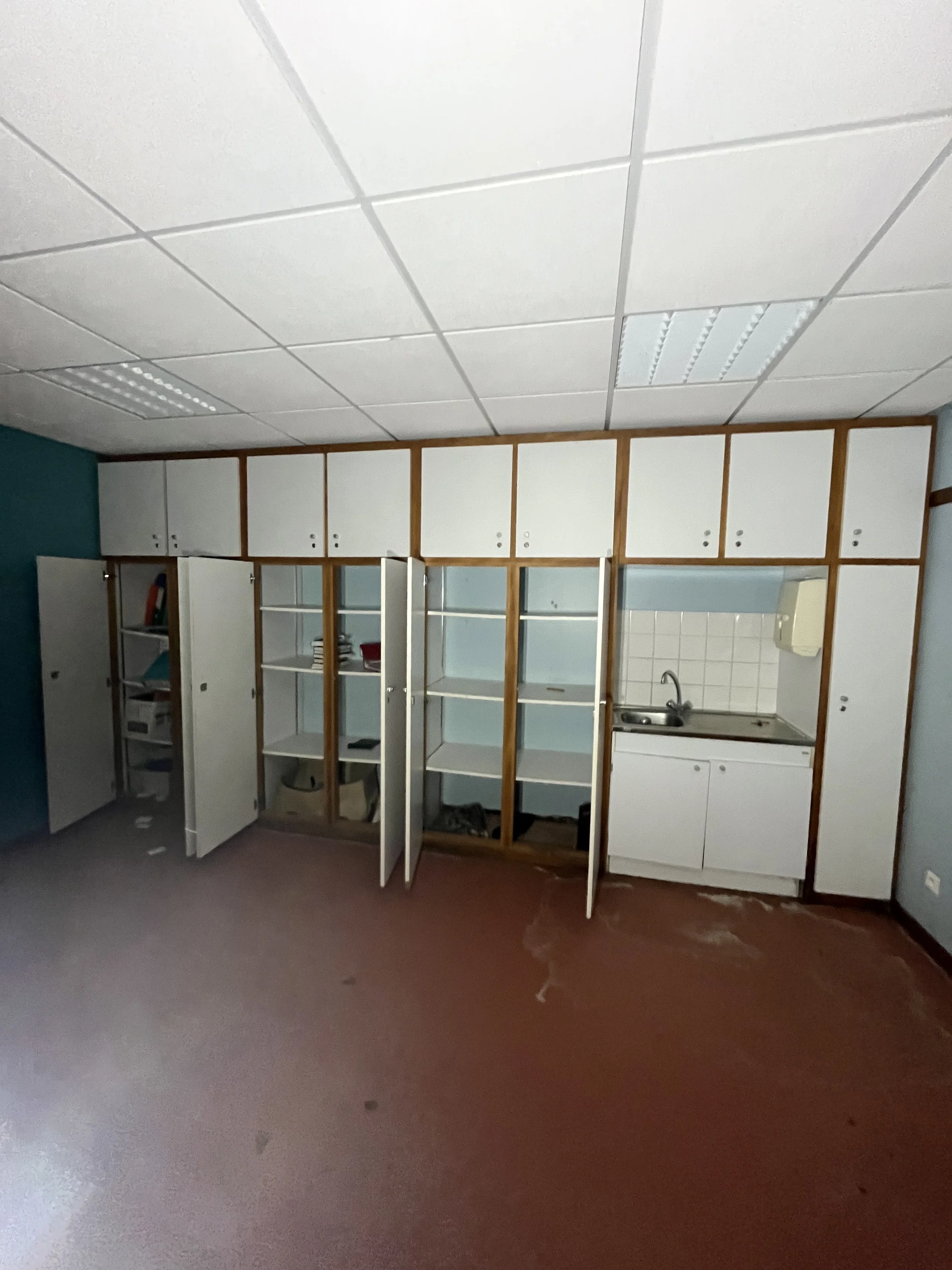 Cuisine avec armoires ouvertes en bois et blanc, évier, et plan de travail, sol rougeâtre, plafond en dalles blanches.