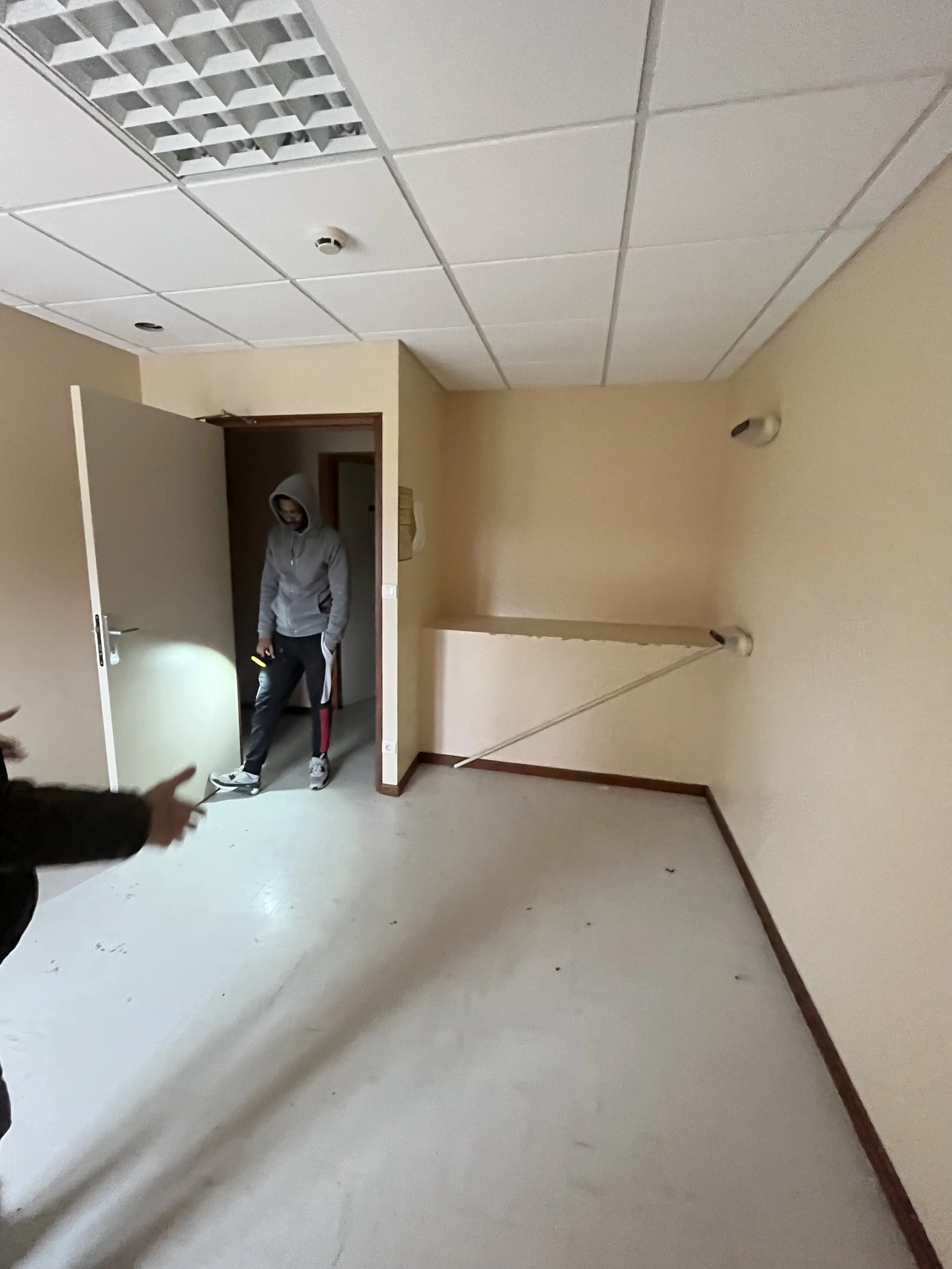 Une pièce vide avec un plafond suspendu, beige mur, une personne avec une capuche gris clair, pantalons noirs et chaussures blanches, debout près d'une porte. La pièce est peu éclairée, il y a une lumière allumée sur la personne et un autre bras poin