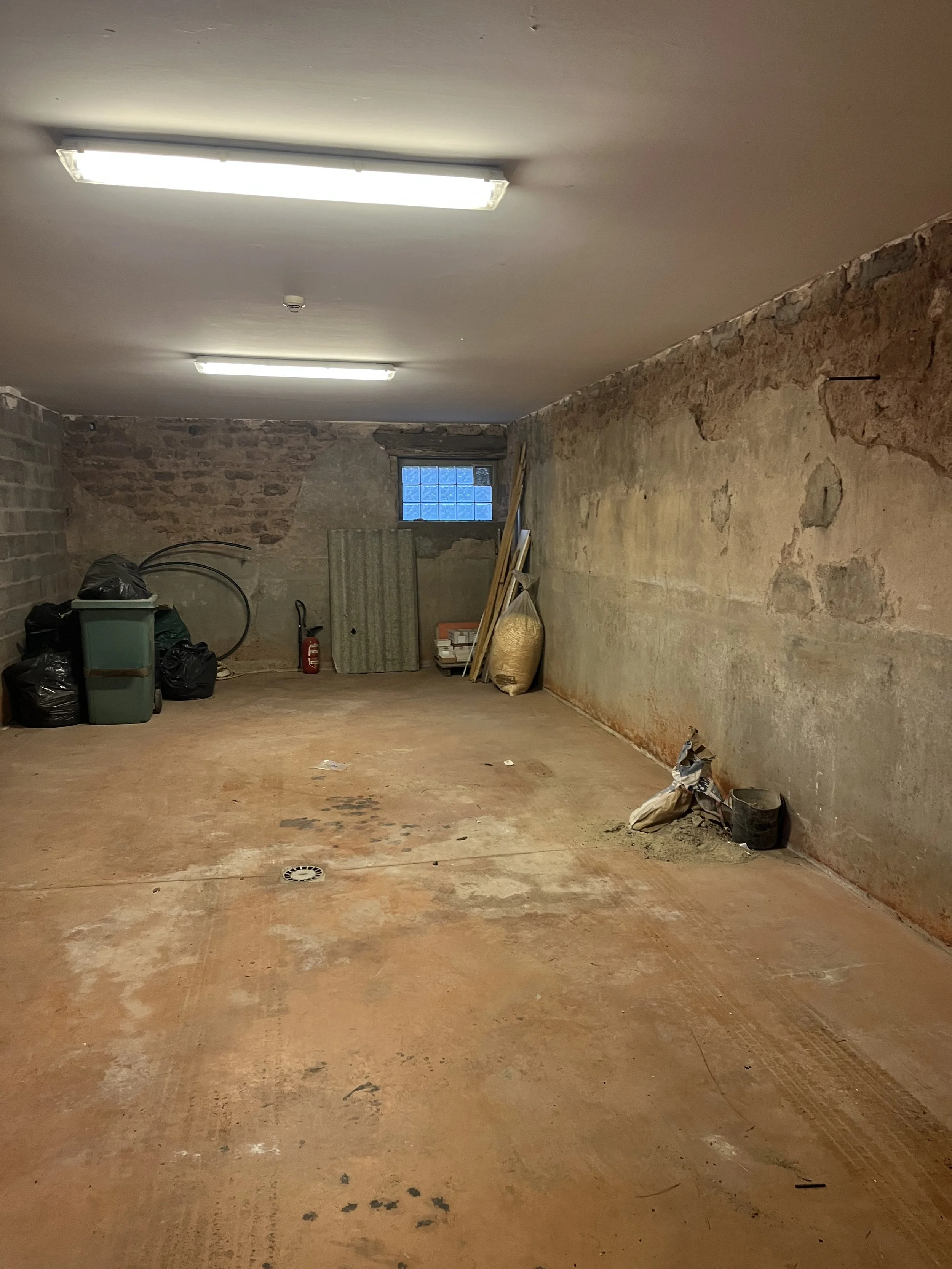 Un sous-sol brut avec murs en pierre et sol en béton, éclairé par des lampes néons, avec des déchets et des outils empilés au fond.