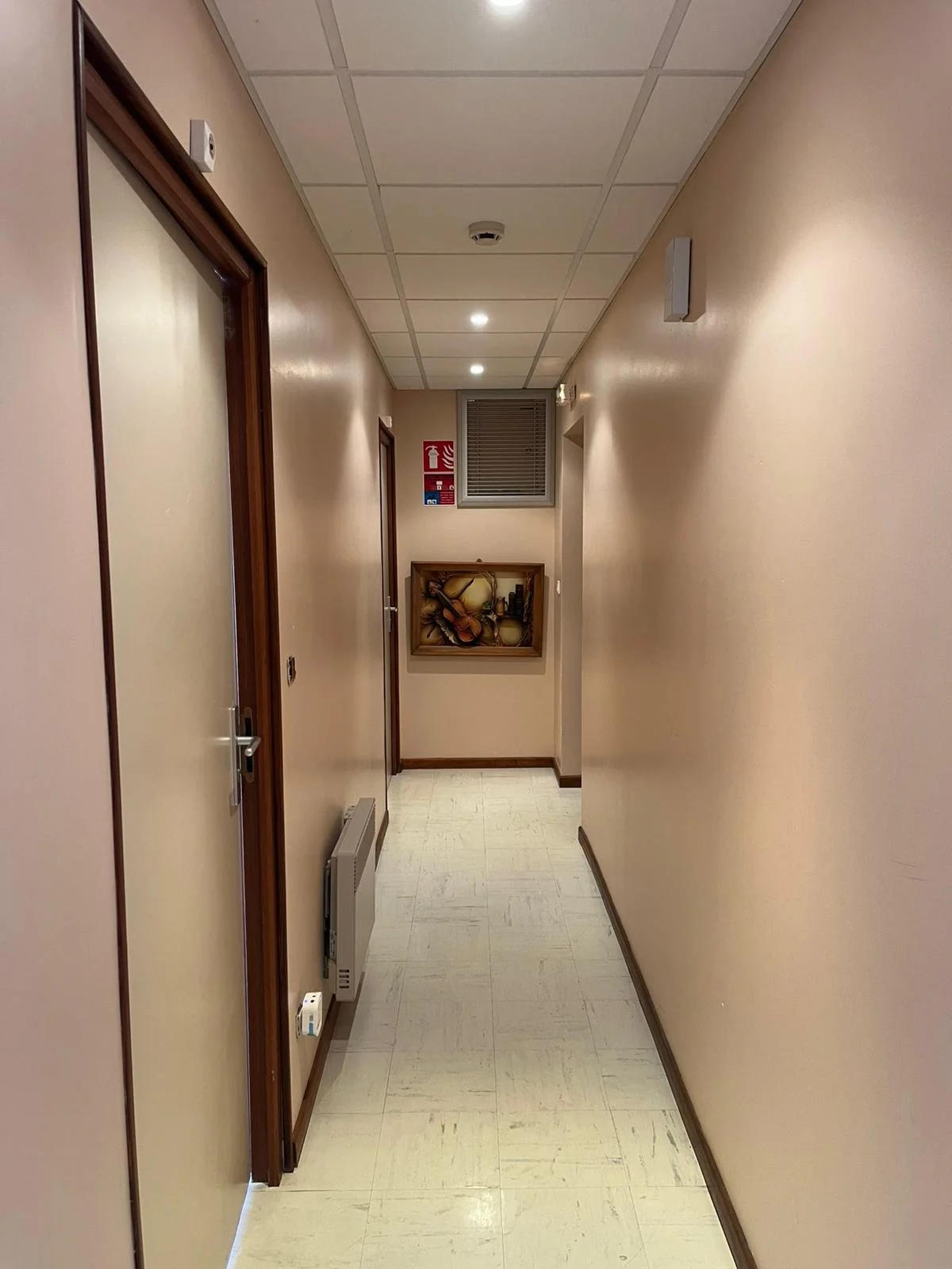 Un couloir intérieur d'un bâtiment avec des murs beige, un plafond en dalles blanches, et un sol en carrelage clair. On voit plusieurs portes en bois sur le côté gauche. À l'extrémité du couloir, il y a une fenêtre et un tableau décoratif avec des in