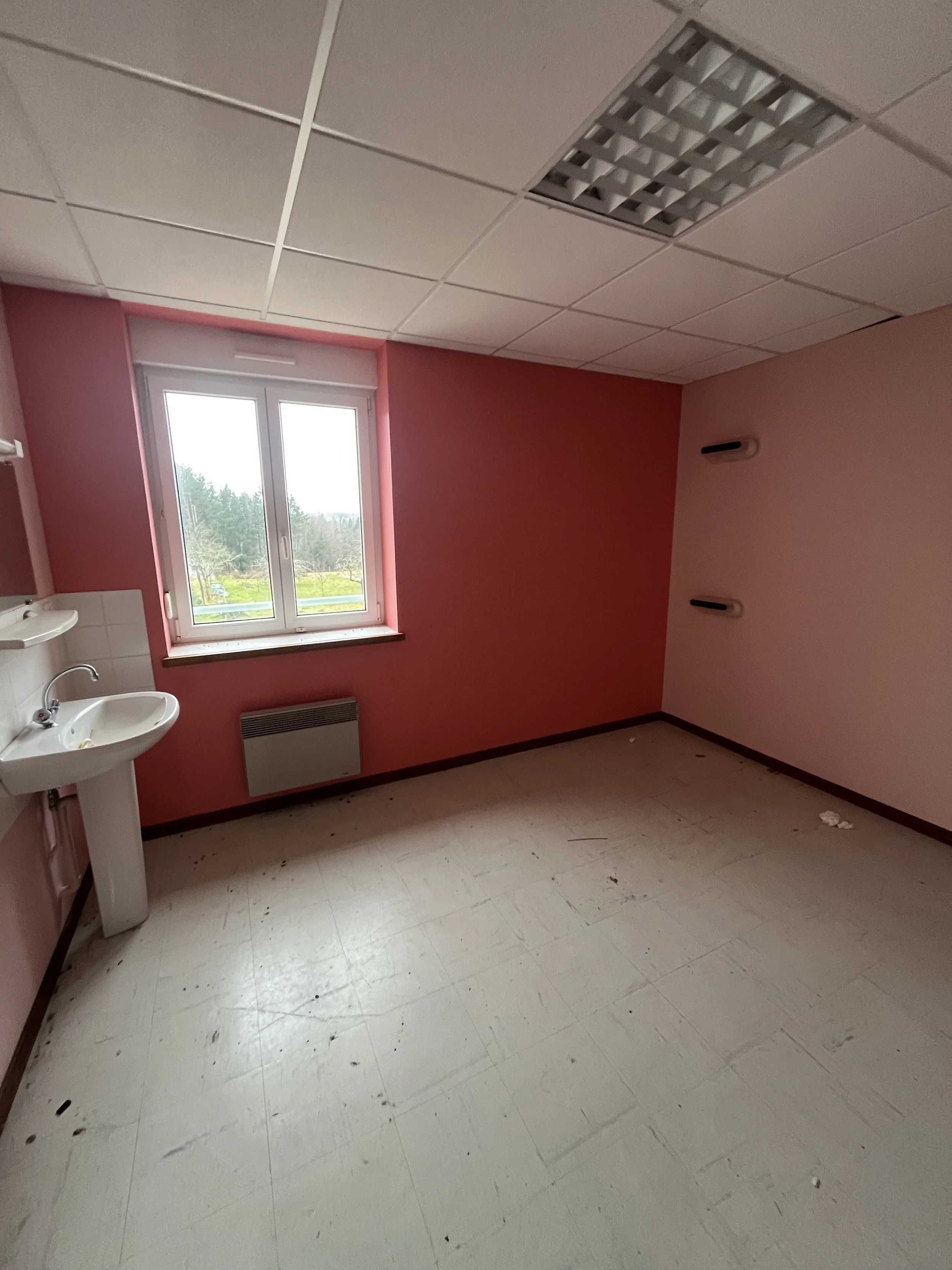 Une pièce vide avec un mur rose, un petit lavabo blanc, une fenêtre, un radiateur, et un plafond suspendu avec des spots lumineux