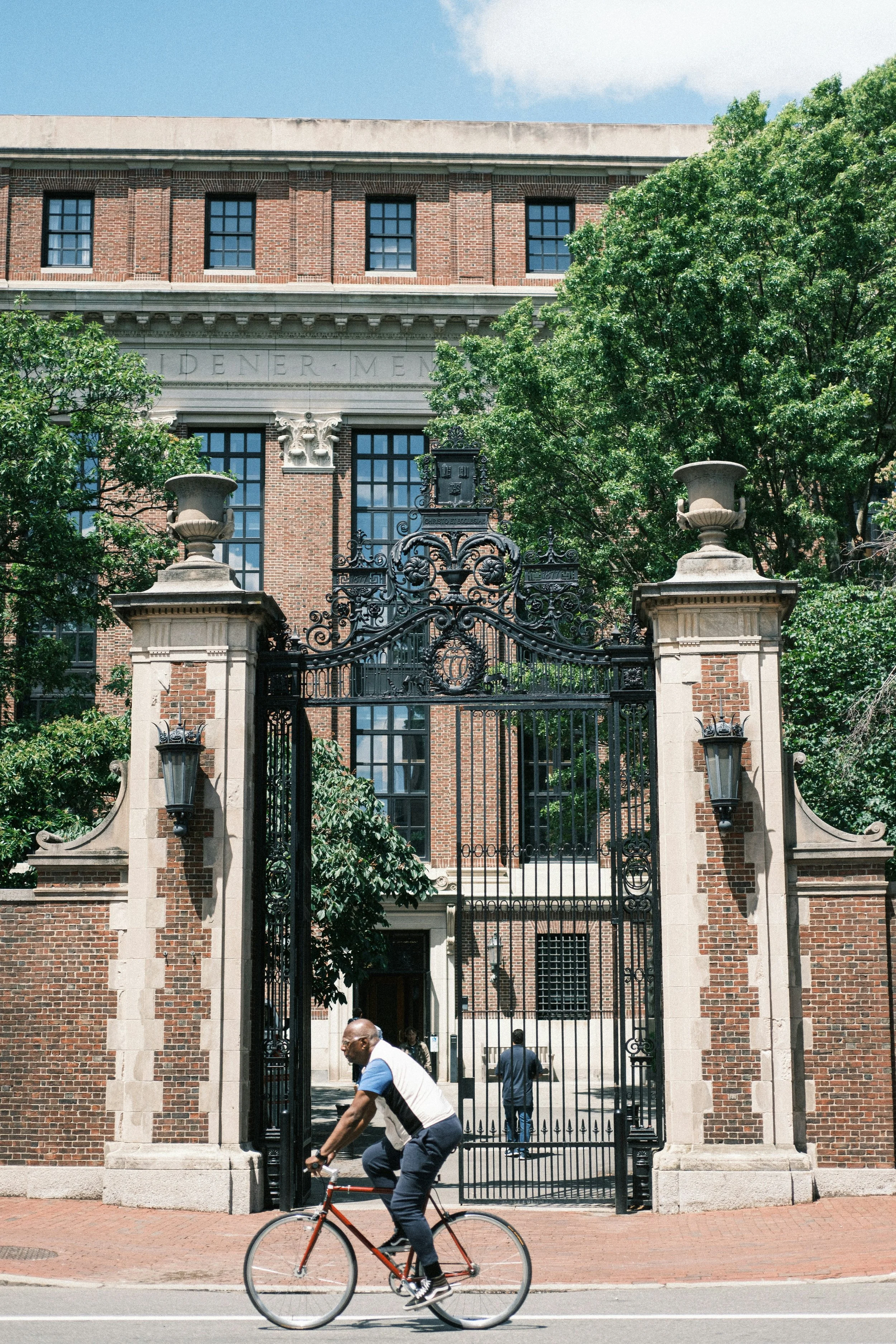 A man riding a bicycle past an ornate iron gate and brick building with green trees.