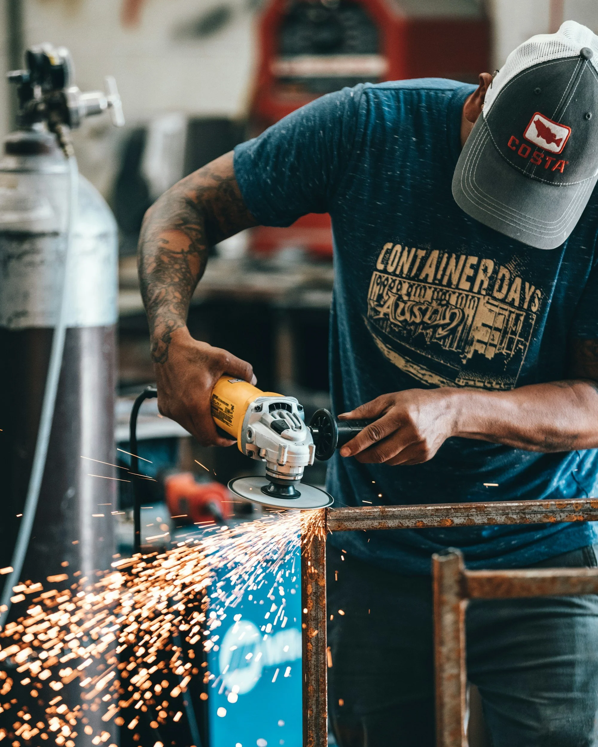 A man wearing a gray cap with a red Costa logo and a blue T-shirt is using a handheld angle grinder to cut a rusty metal pipe, creating sparks in a workshop or industrial setting.