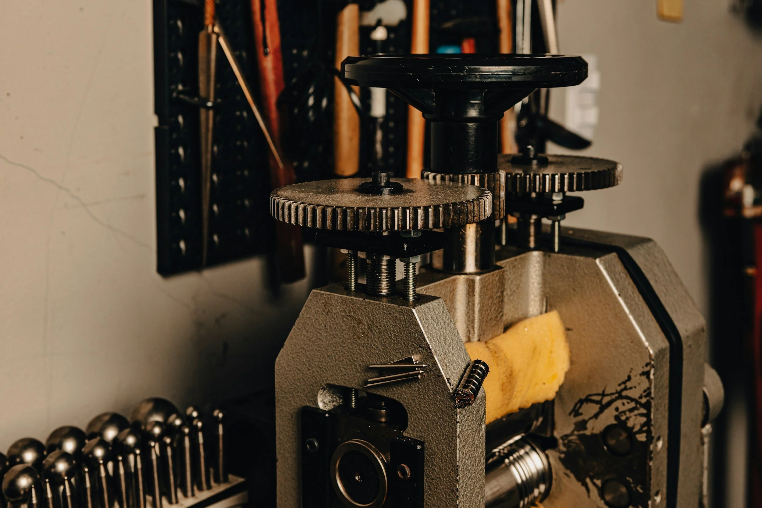 Close-up of a mechanical device with gears, screws, and metal parts on a workbench, with tools and equipment in the background.