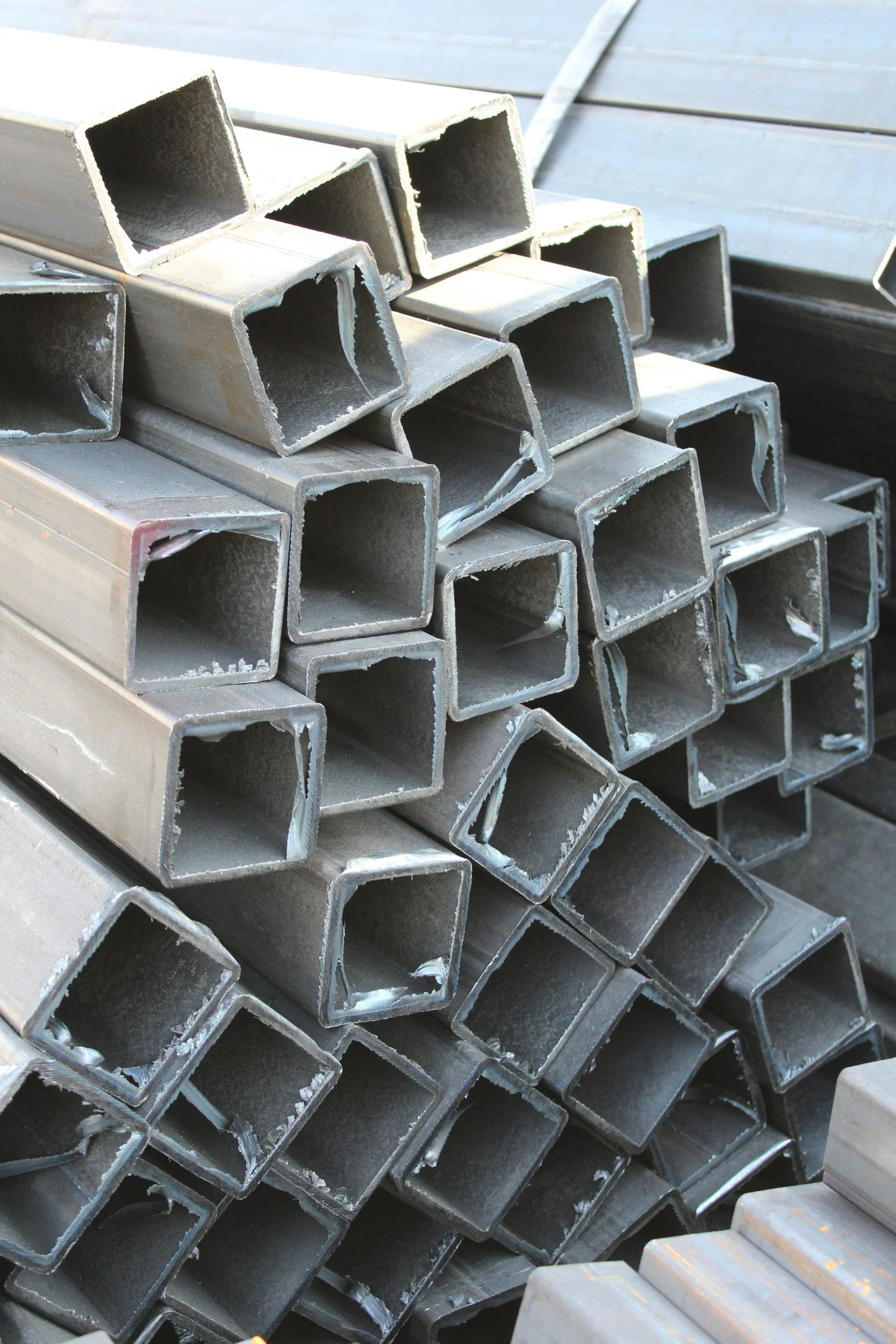 Stack of metal square tubes, some with rough edges, outdoors.