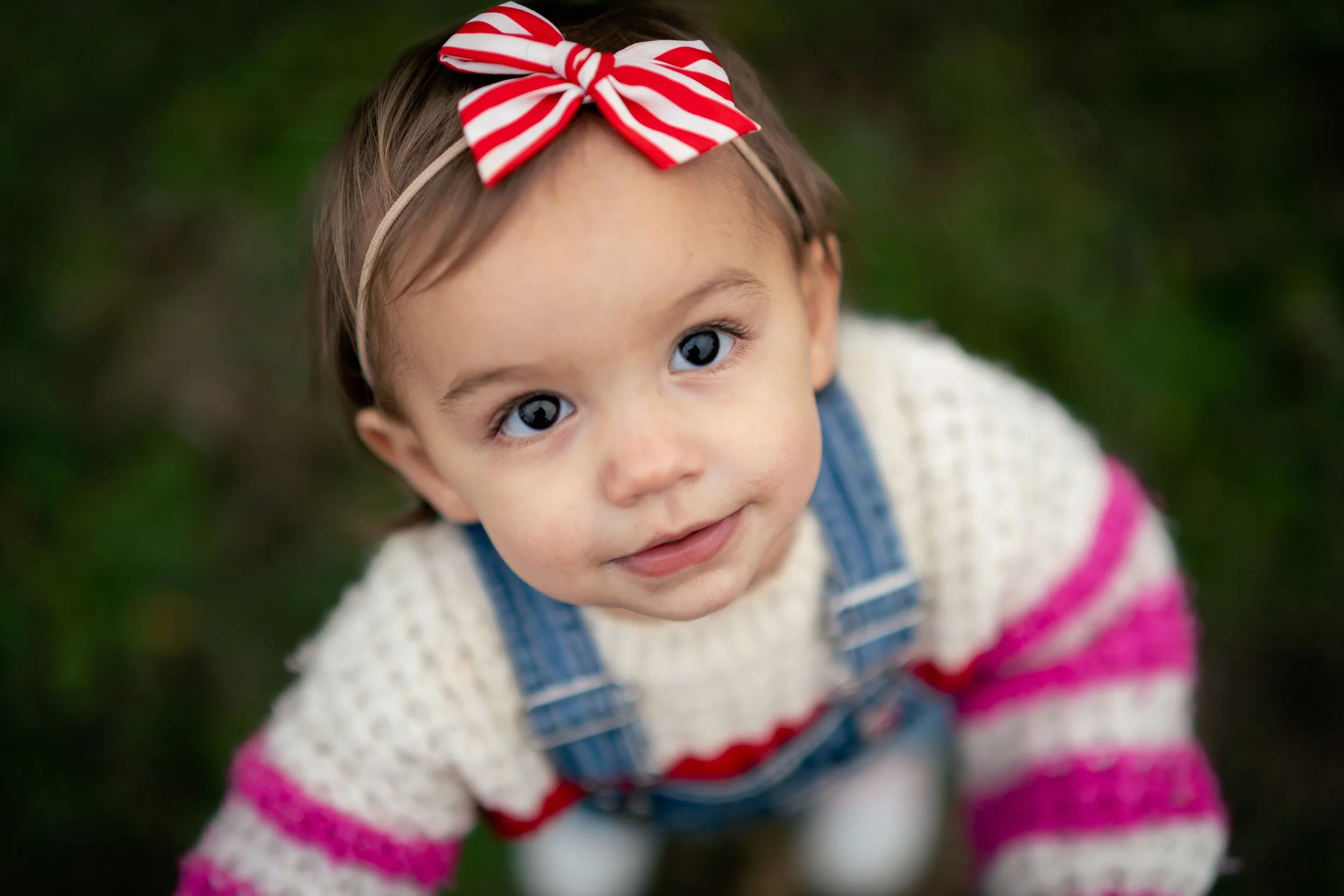 A young girl with big blue eyes, wearing a red and white striped bow headband and colorful striped sweater, looking up at the camera outdoors in North Alabama