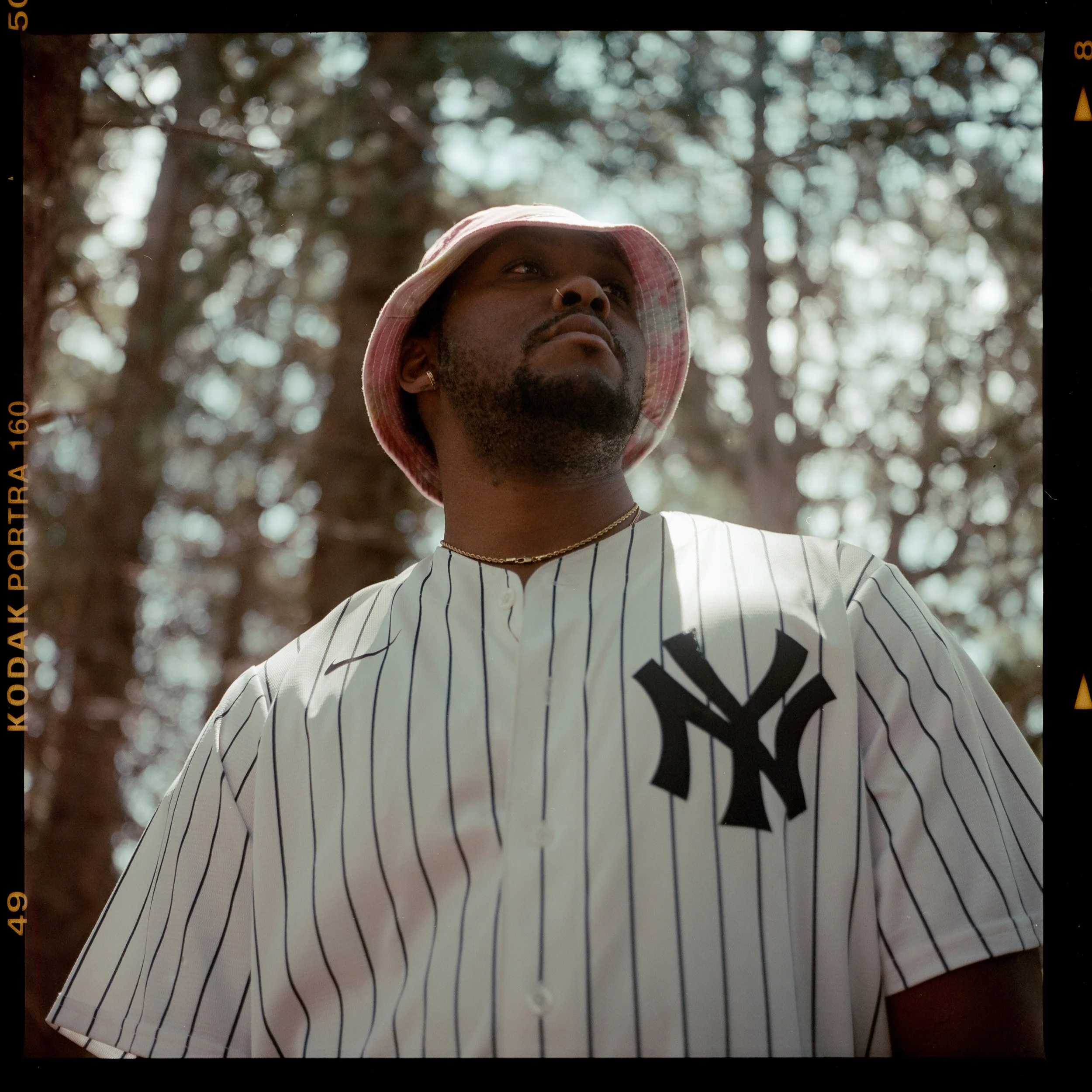 Film photography capturing man wearing a New York Yankees baseball jersey, a pink bucket hat, and a gold chain, standing outdoors in a wooded area with trees in the background in Hamilton, Alabama