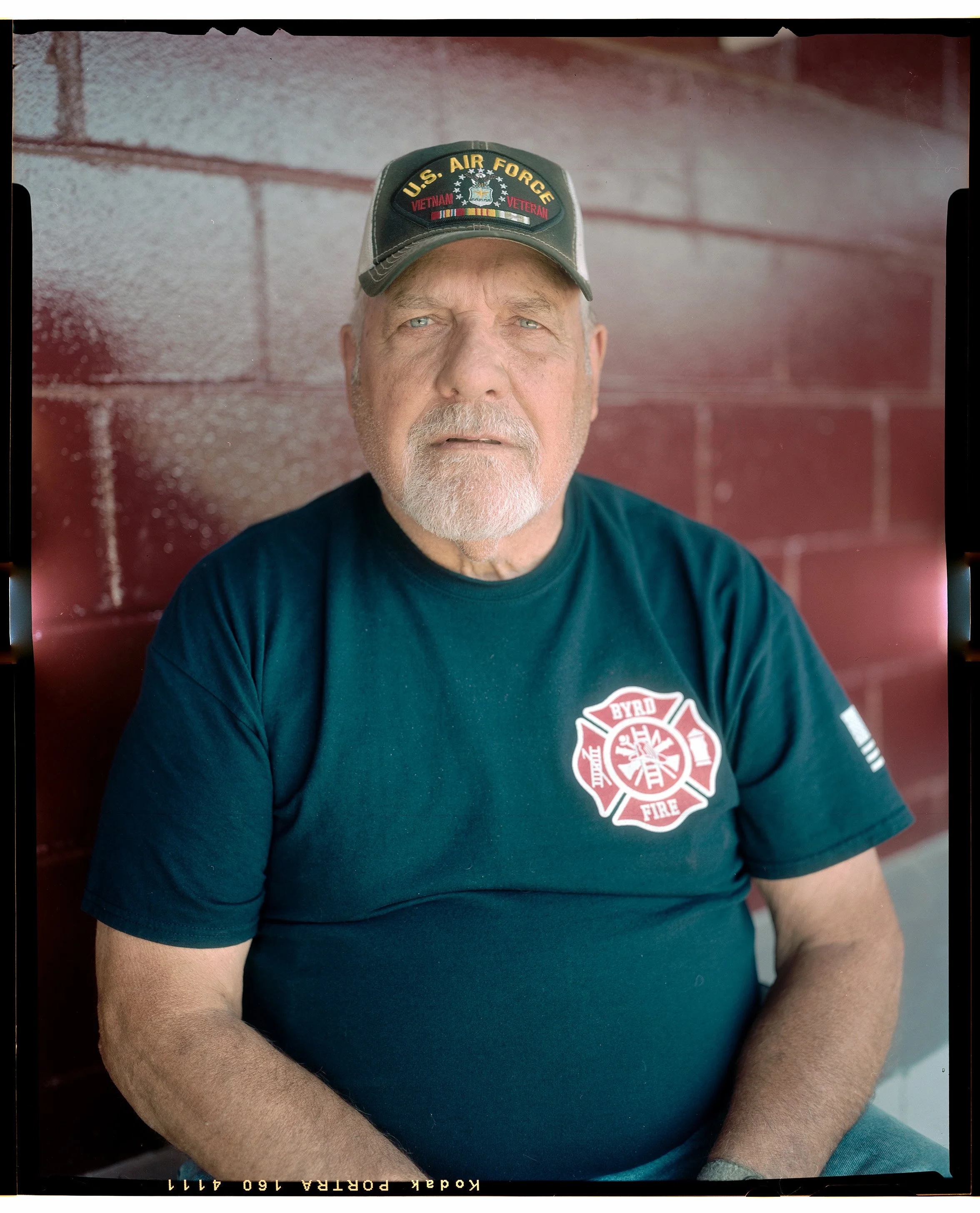 A Northwest Alabama man with a white beard and blue eyes wearing a dark green U.S. Air Force Vietnam veteran cap and a black T-shirt with a red and white fire department emblem, sitting against a red brick wall.