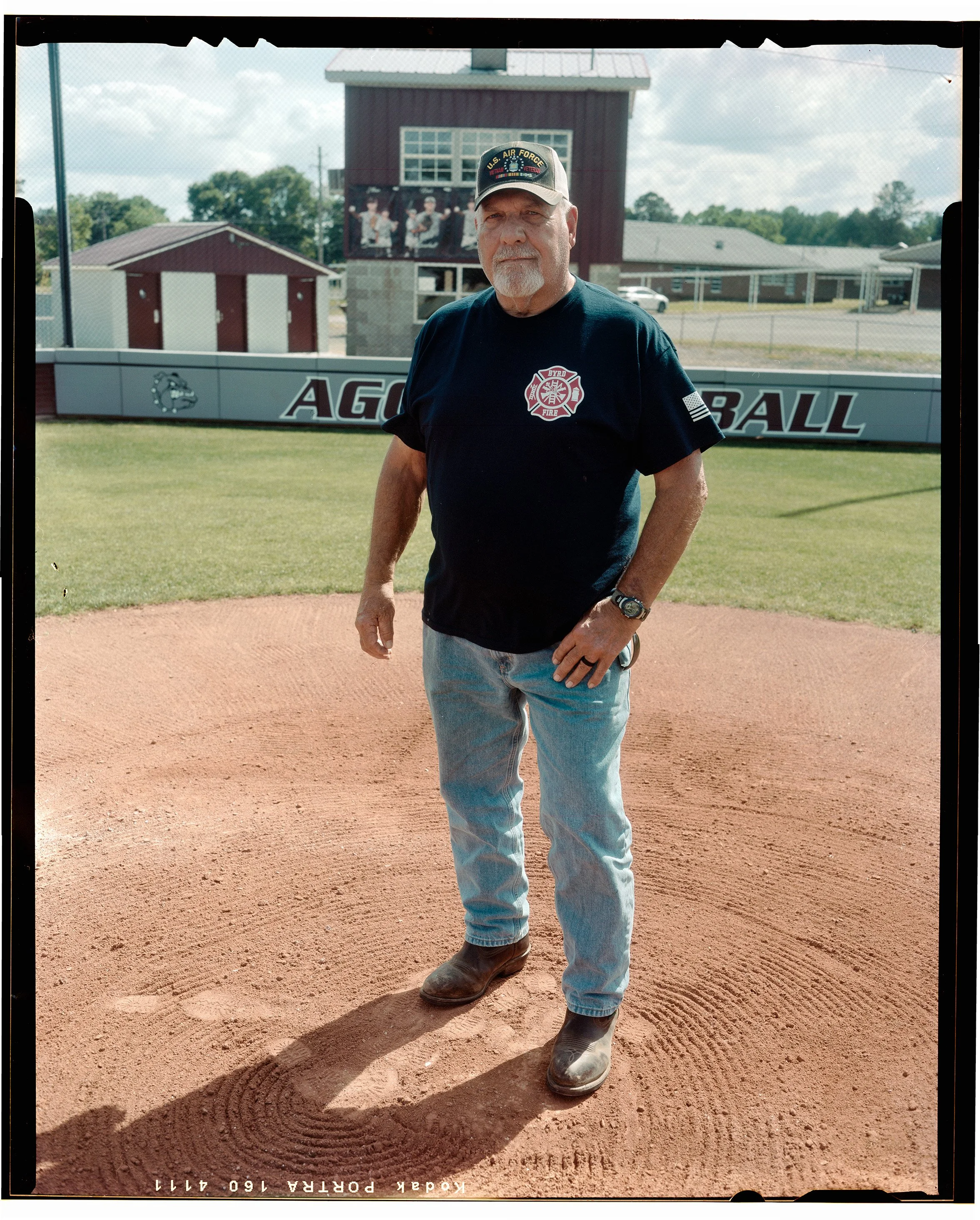 A Hamilton, Alabama man standing on a baseball pitcher's mound at a baseball field, wearing a U.S. Air Force cap, a navy fire department t-shirt, jeans, and cowboy boots.