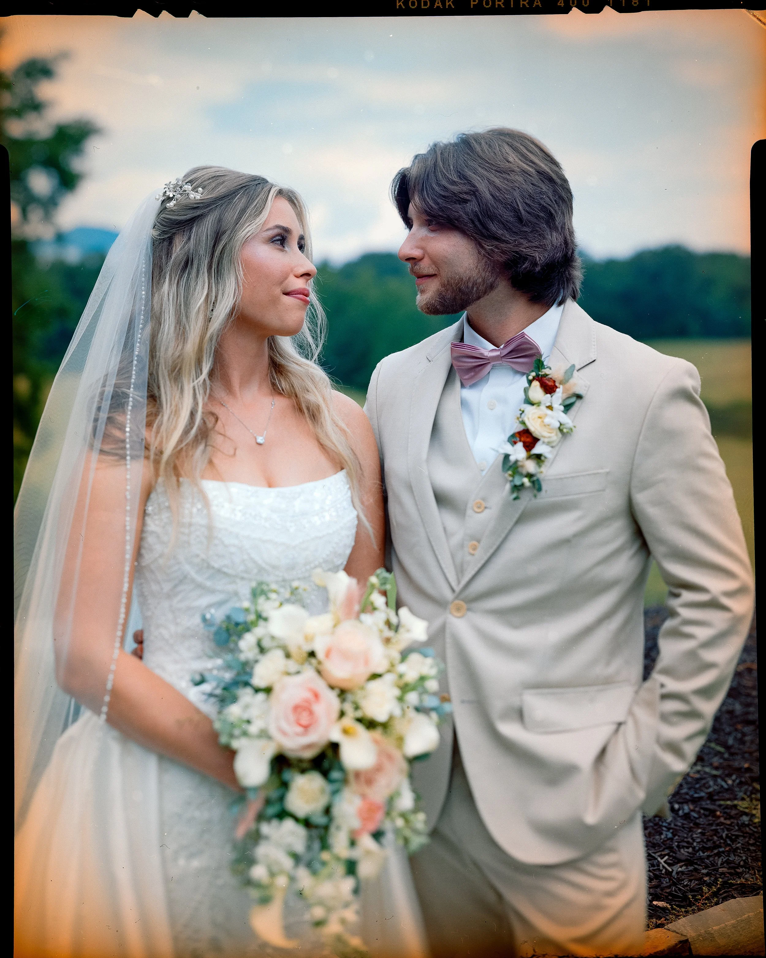 A bride and groom standing outdoors at sunset, looking at each other. The bride is wearing a white wedding dress with a veil and holding a bouquet of roses and white flowers. The groom is dressed in a light-colored suit with a pink bow tie and a bout