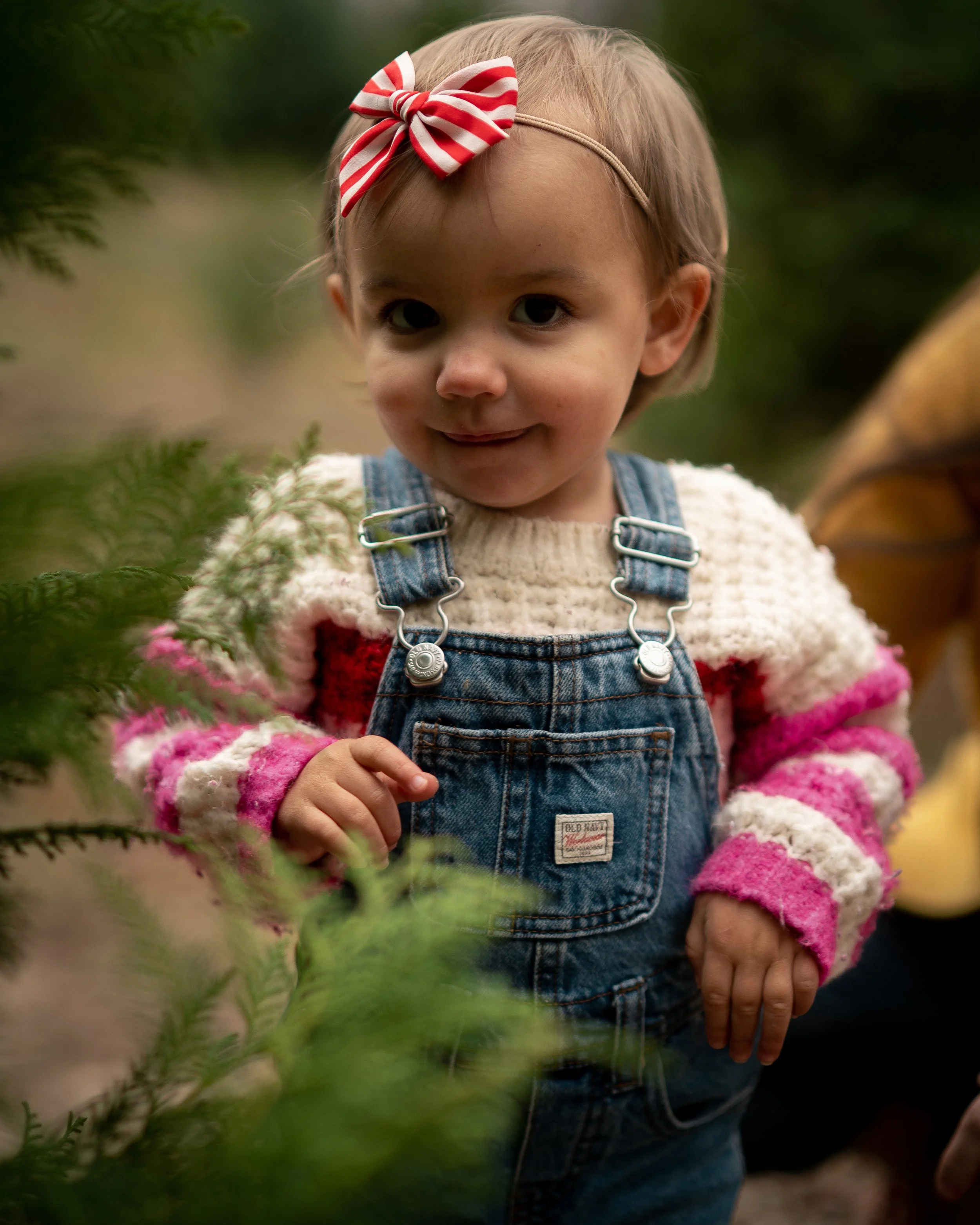 A young girl with a red and white striped bow headband, wearing a cozy, colorful sweater and denim overalls, standing outdoors at the Christmas Tree Farm in Alabama