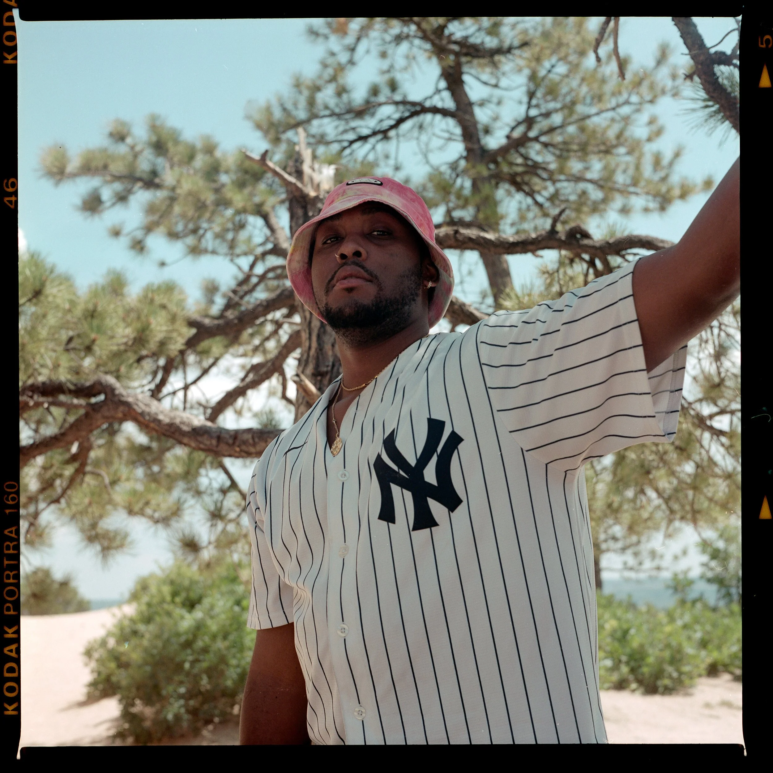 Film photography capturing a man utdoors on a sunny day, wearing a white and navy striped New York Yankees jersey, a pink bucket hat, and gold jewelry, with green trees and a clear sky in the background in Hamilton, Alabama