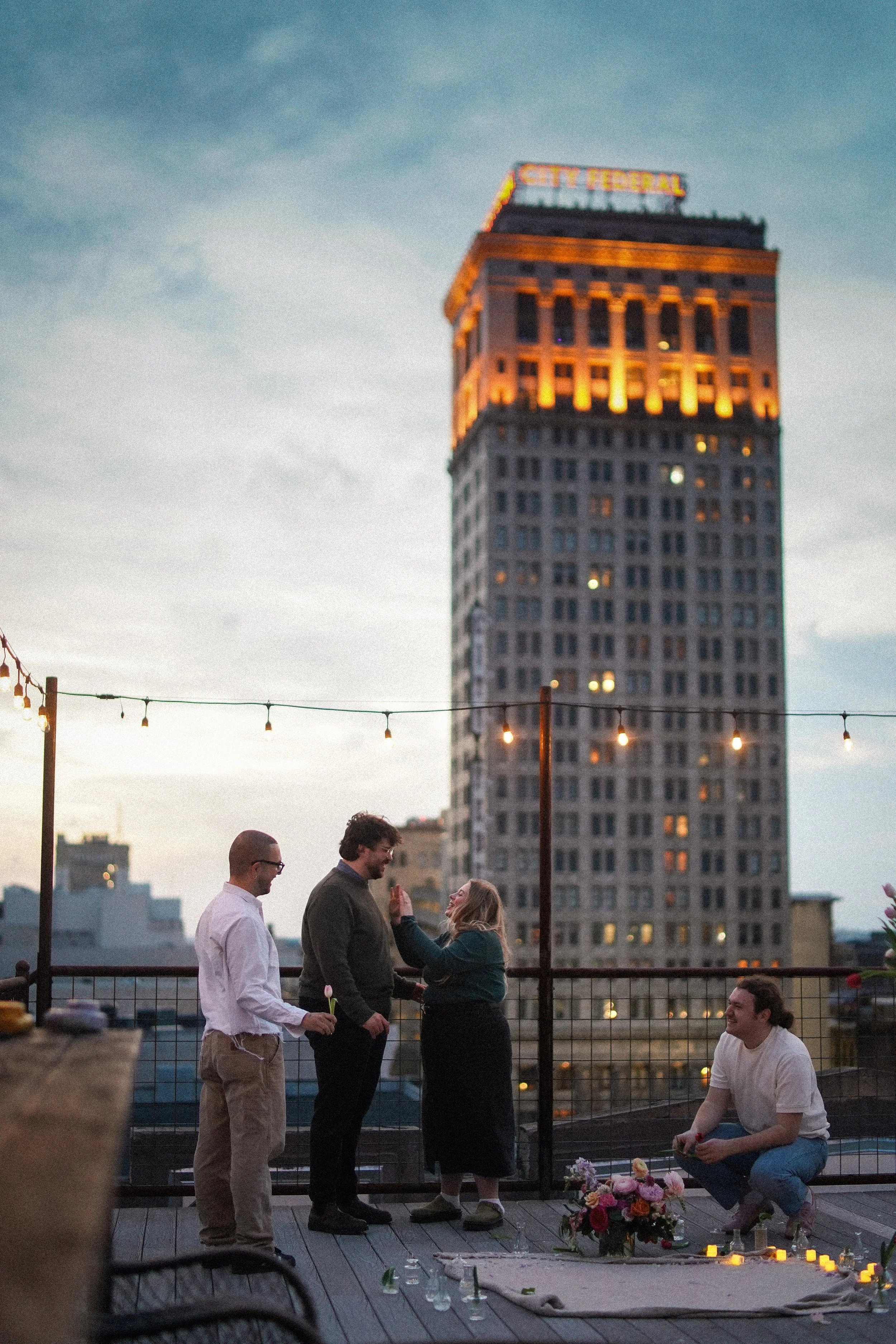 People gathering on a rooftop at dusk, with a city skyline and lit-up skyscraper in the background, engaged in conversation and romantic gestures.