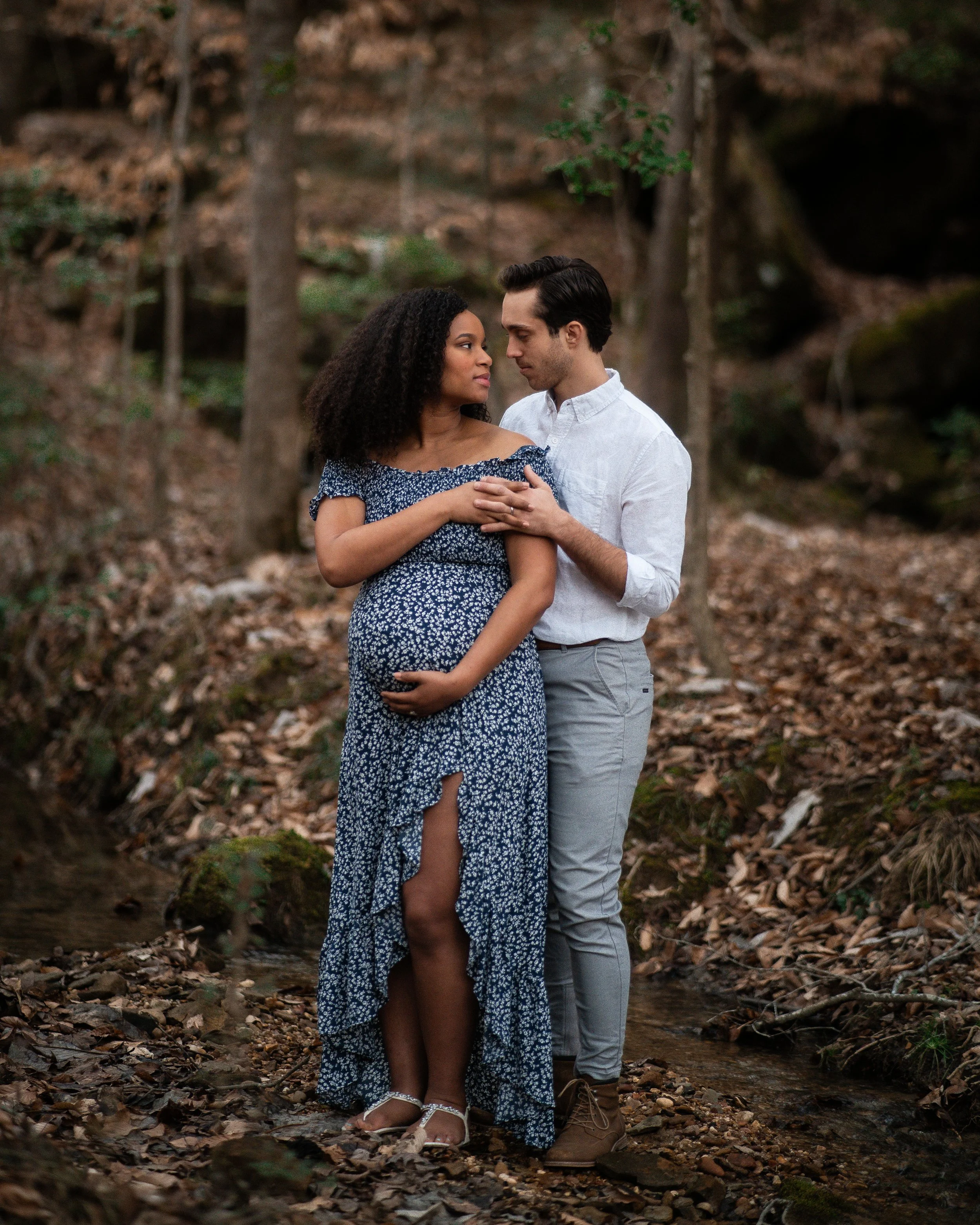 A pregnant woman in a blue floral dress and a man in a white shirt and gray pants standing close together in a forest, holding hands and looking into each other's eyes in North Alabama