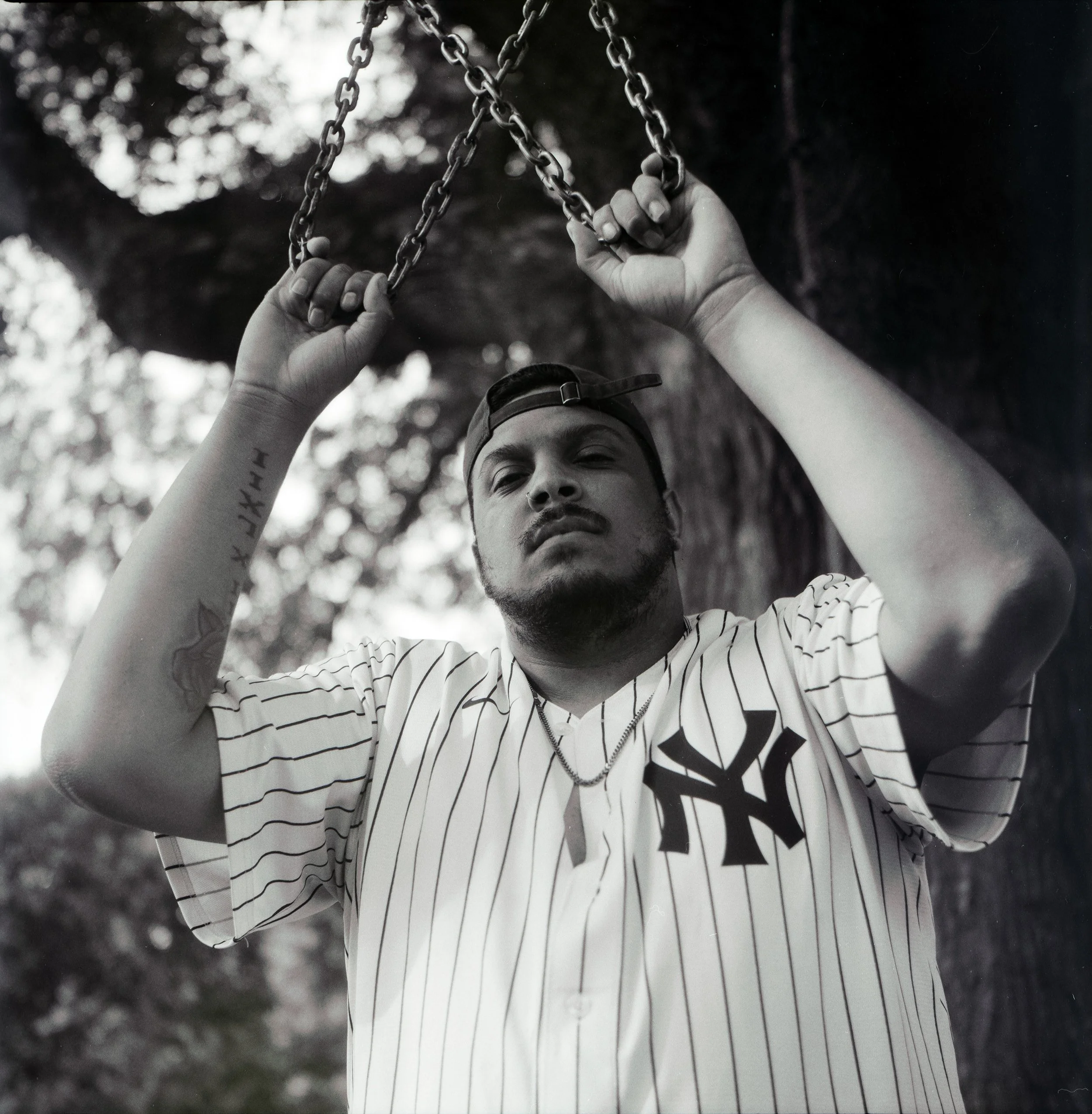 Black and white photo of a man wearing a baseball cap backwards and a New York Yankees baseball jersey, holding onto chains hanging from above, outdoors with blurred trees in the background.