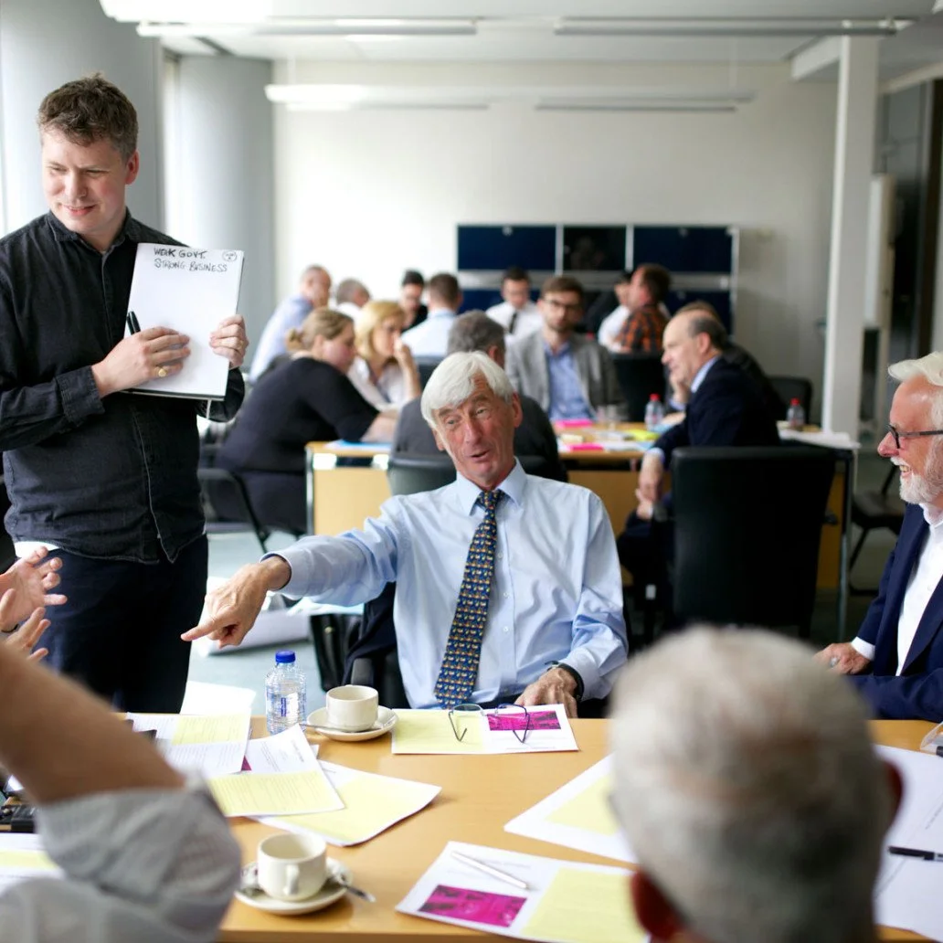 People in a meeting room engaged in a discussion. One man is pointing while others listen; some are taking notes. A young man is holding a notebook with the words 'Work Gov't Shaping Business' written on it.